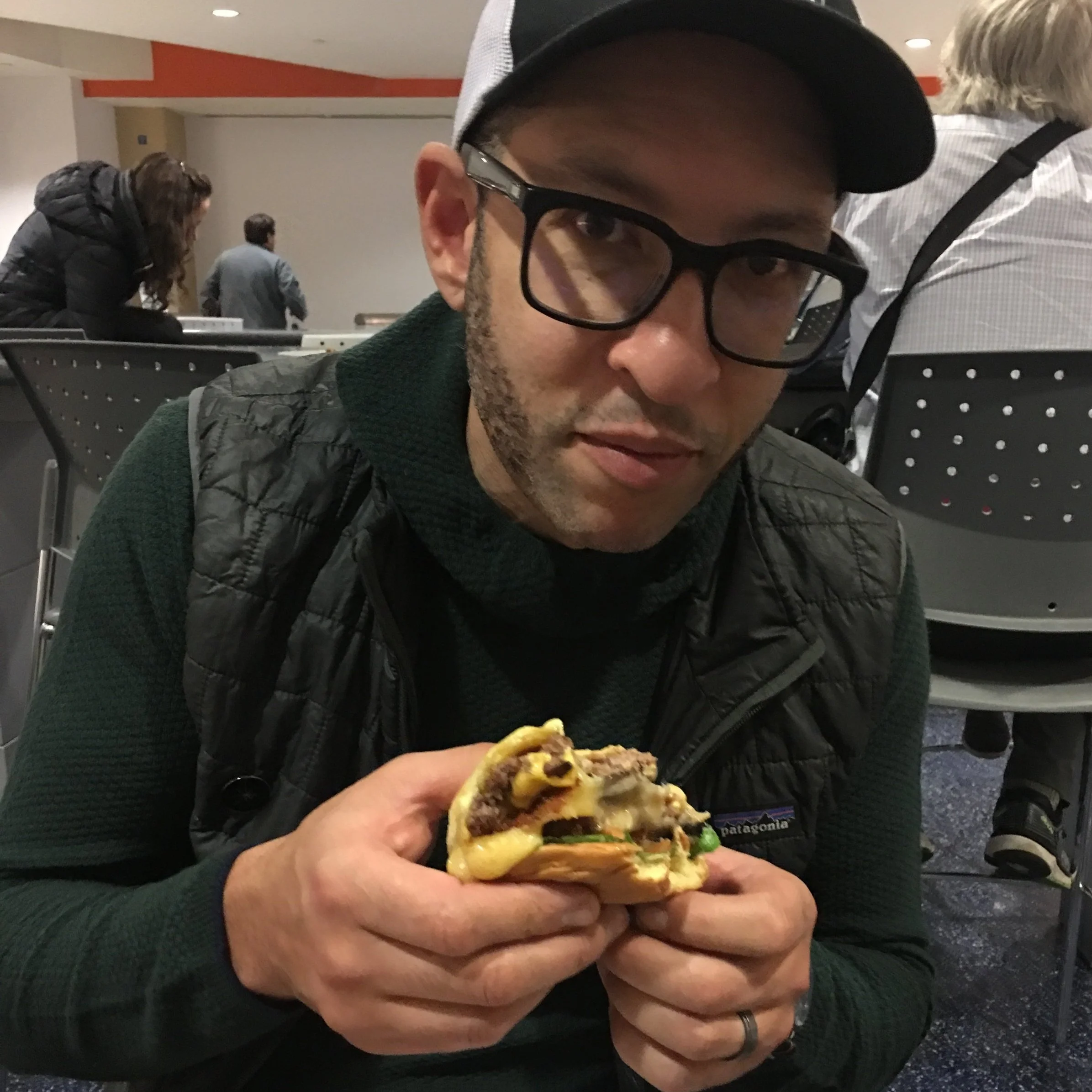 Sean Randall pictured holding a large burger with a few bites missing. He has a slight smile while looking at the camera. He is wearing dark casual clothes, a ball cap, dark rimmed glasses, and has his beard closely cropped.