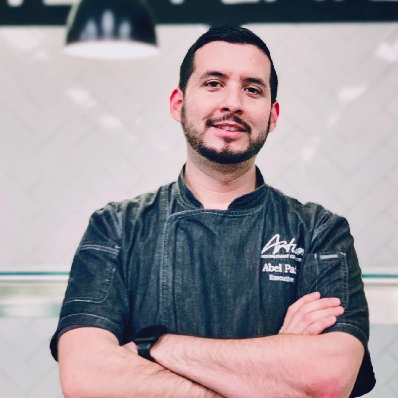 Abel Padilla has short dark hair and close cropped beard, is wearing a dark chefs coat with short sleeves. His arms are crossed and he is softly smiling.