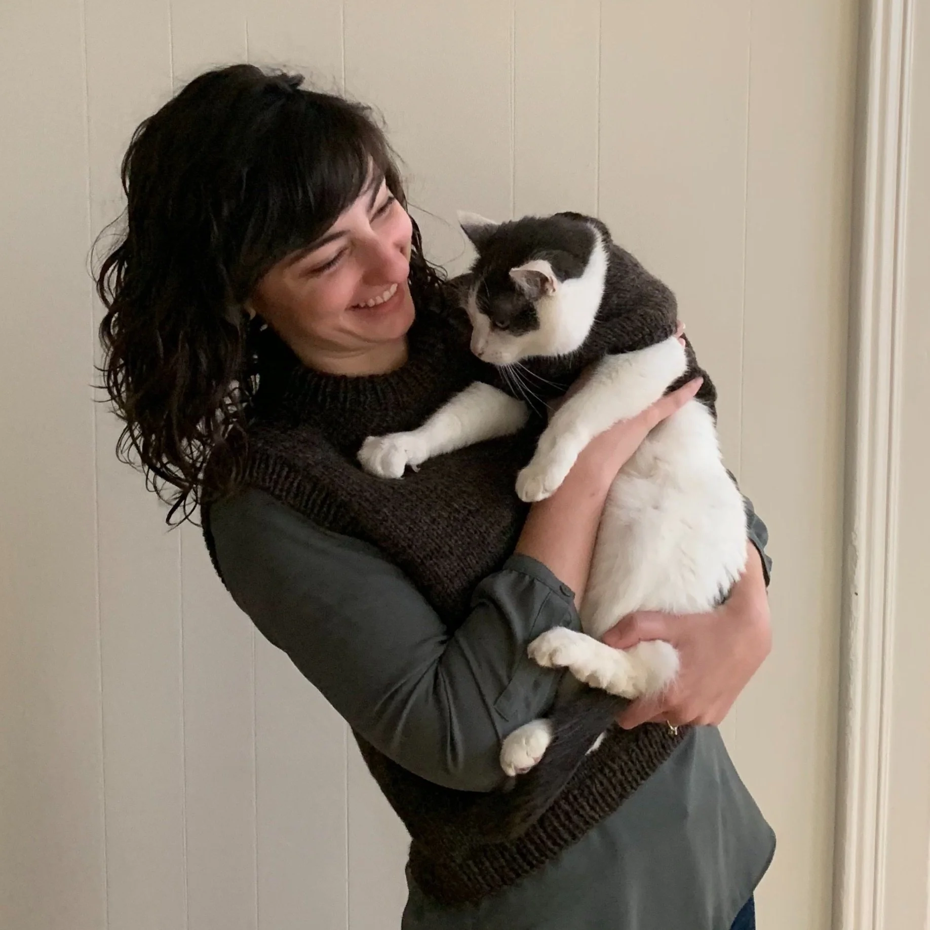 Leslie Eckert-Page pictured with one of her cats, they are wearing matching brown knitted sweaters. Leslie is holding the cat and smiling.