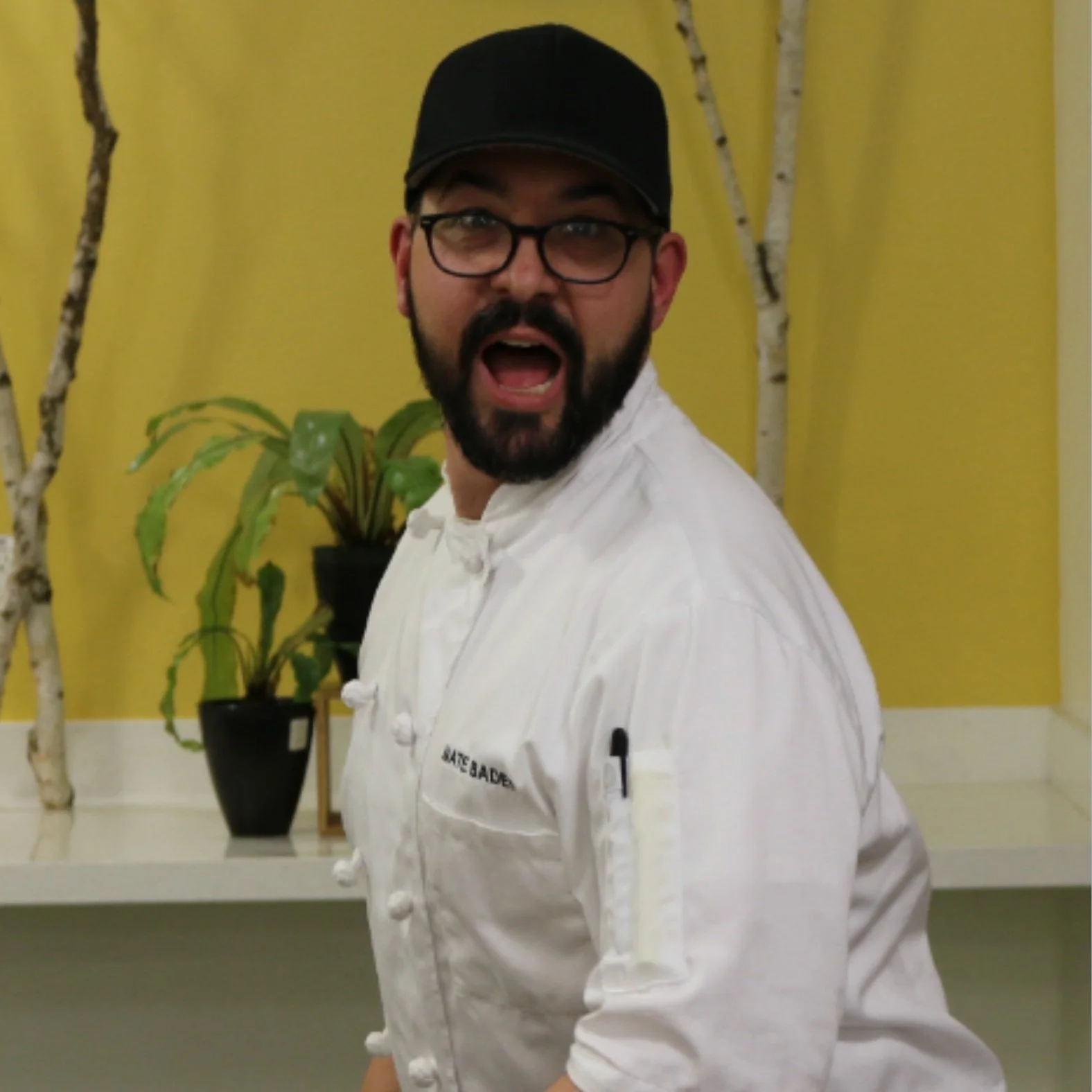 Nate Bader caught middle of turning to run, he is wearing a white chefs coat, black ball cap, and dark rimmed glasses with closely cropped dark beard. There are potted plants and a yellow wall in background.