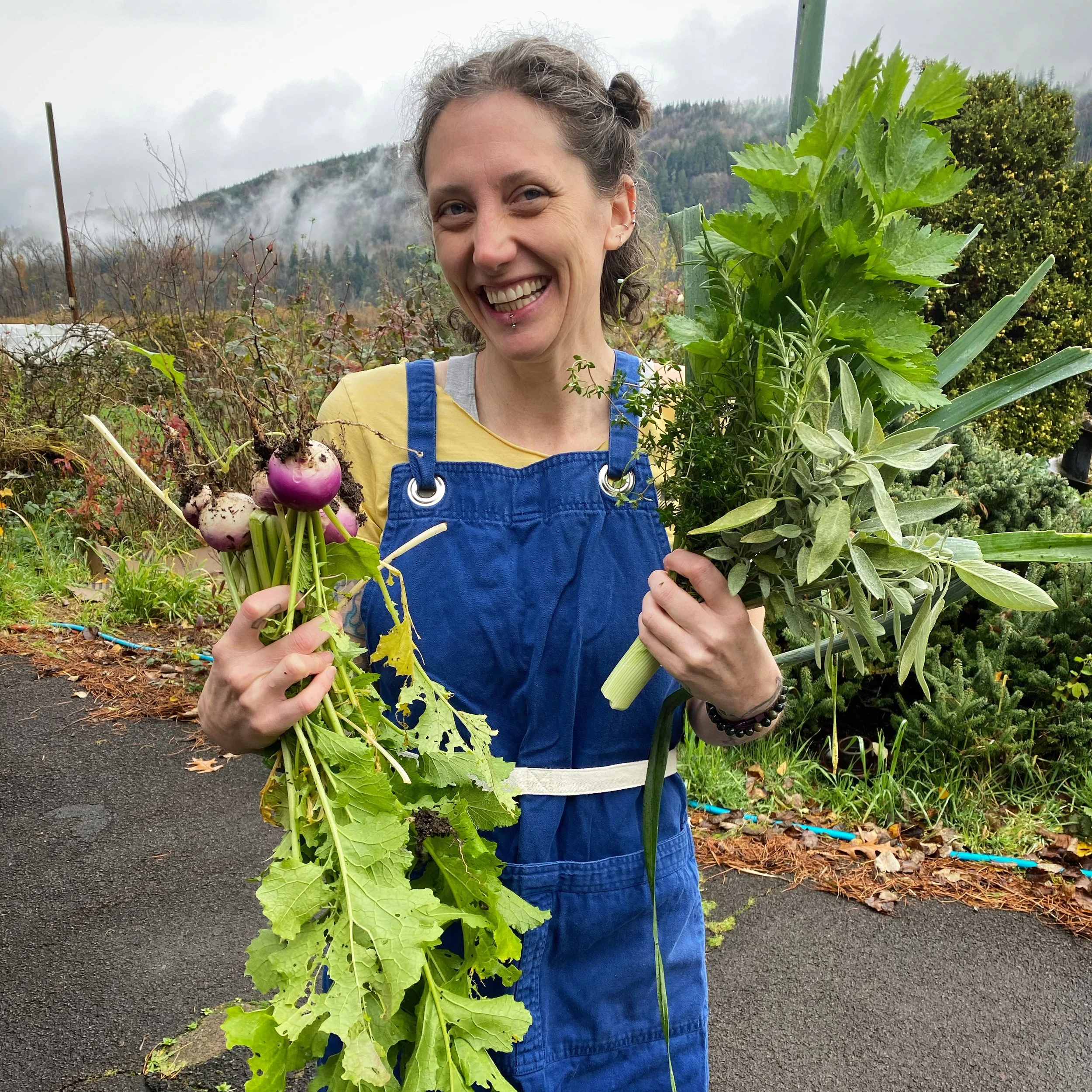 hillary lauren smiles widely while holding freshly harvested purple turnips, leeks, and a variety of herbs. She’s in the garden on a partly cloudy day wearing a blue apron over a yellow shirt. (hillary lauren is formerly known as Hillary L. Bergh)