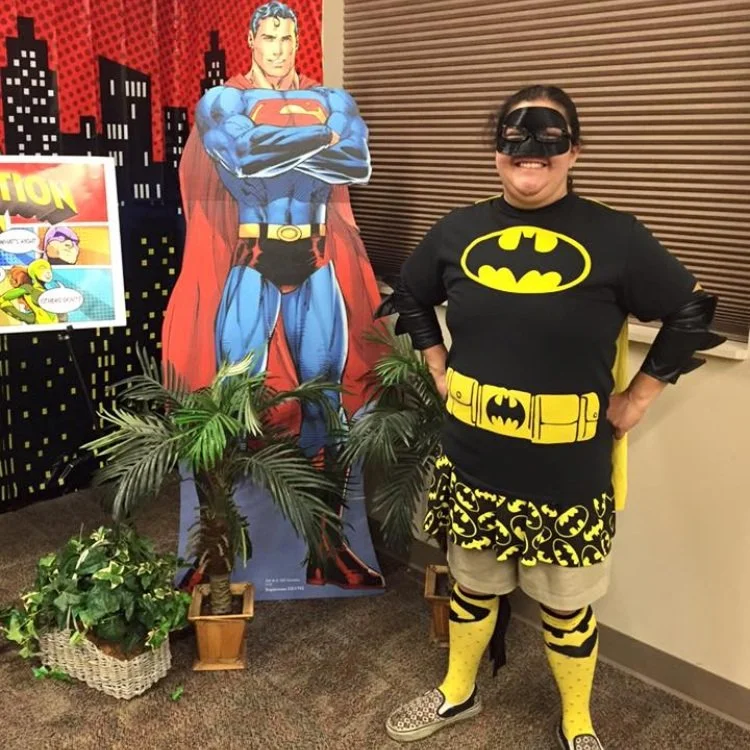 It's Batman! No, it's Cynthia Resendiz in a black and yellow Batman costume and face mask. Cynthia is standing in superhero pose and there is a cutout of Superman in the background.