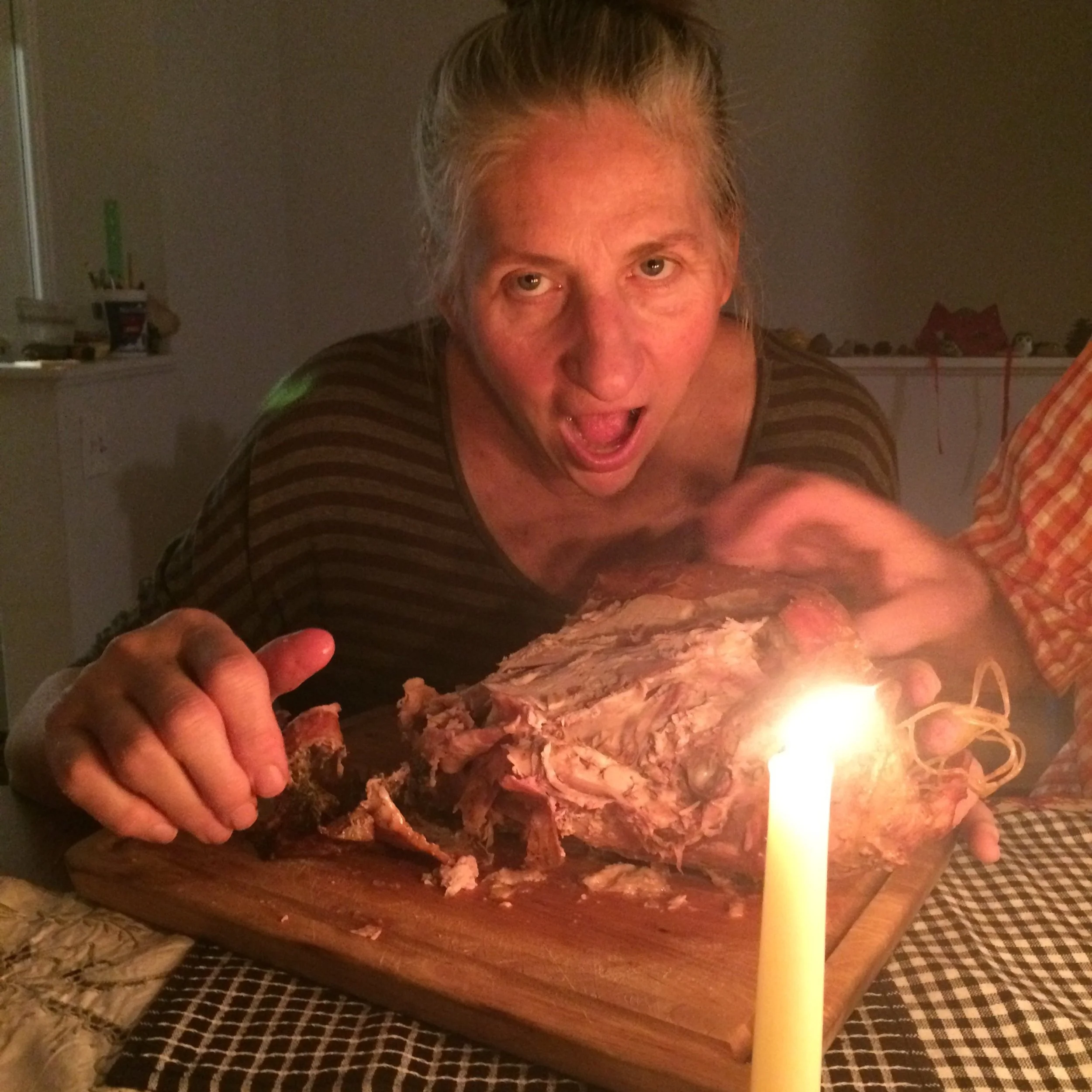 JanyRae (Janet) Seda with a large turkey carcass after Thanksgiving, she is posed to take a big bite. Her salt and pepper hair is pulled back from her face and there is a candle in the foreground.