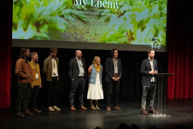 Group of six people standing on stage in front of a large screen, one person speaking at a podium.