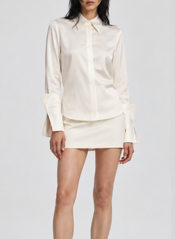 A woman wearing a cream-colored silky blouse with long sleeves and a matching cream-colored mini skirt, standing against a plain background.