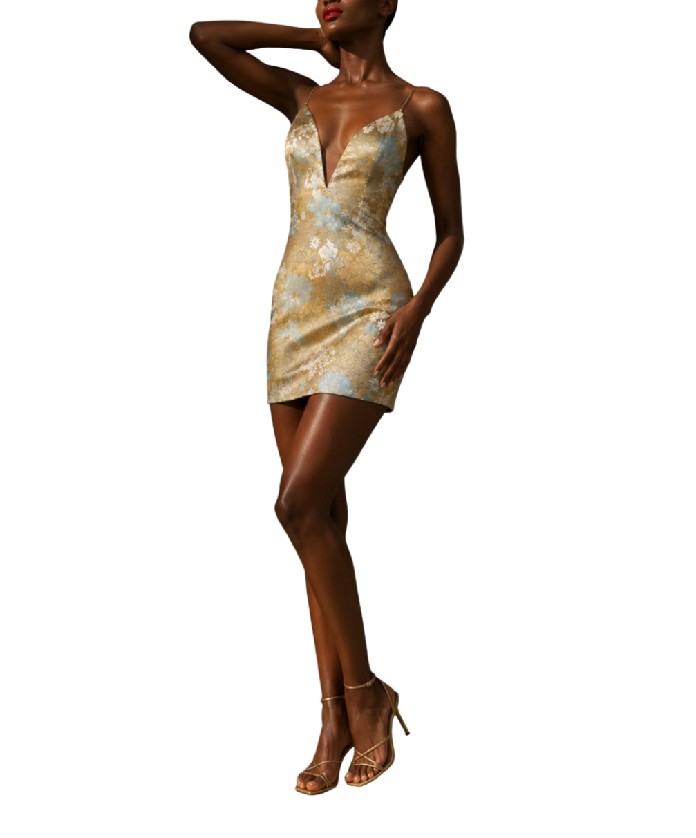 A woman wearing a gold and light blue floral-patterned short slip dress with spaghetti straps, standing with one hand on her neck and the other on her thigh, wearing high-heeled sandals.