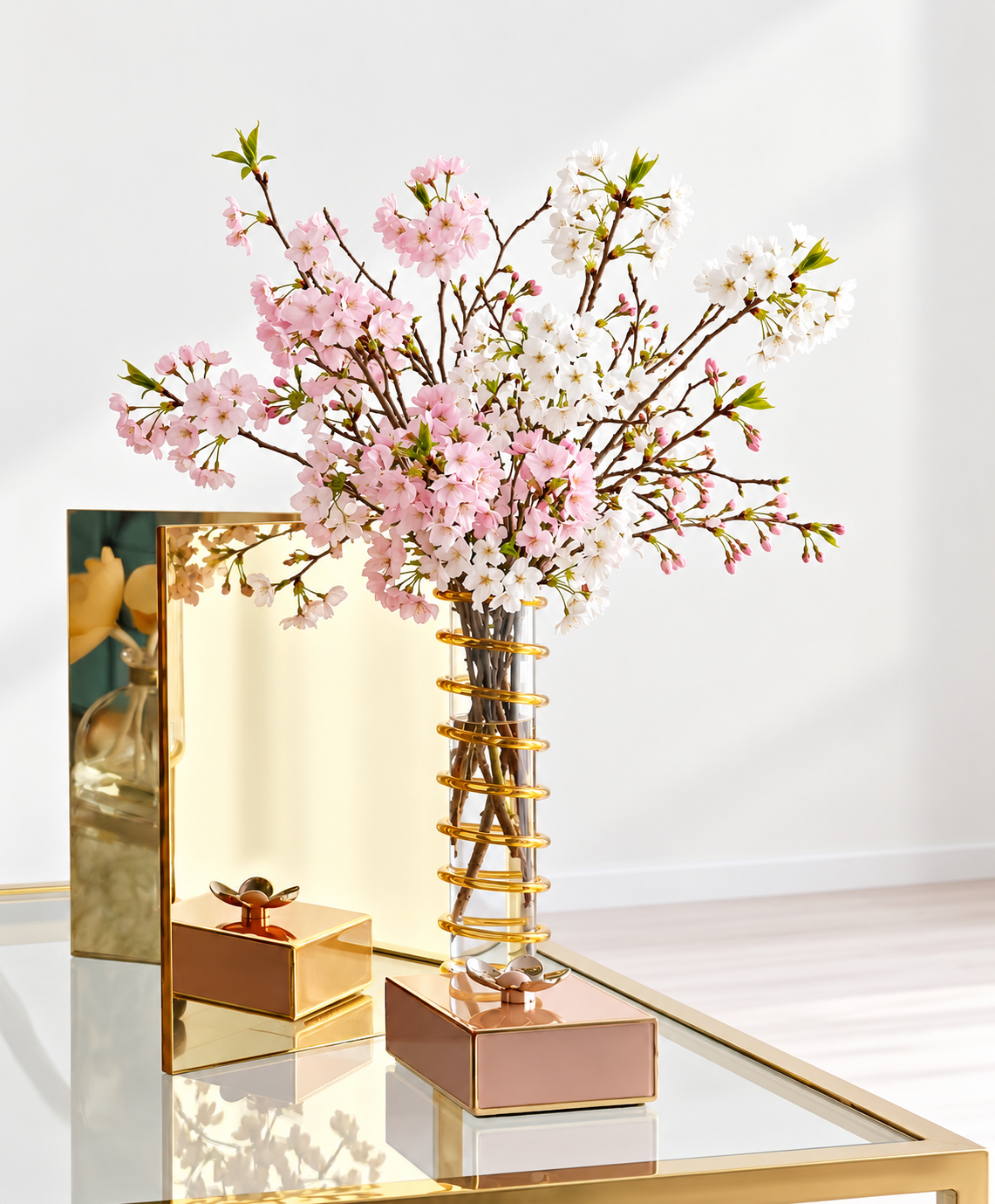 A tall glass vase with pink and white cherry blossom branches on a glossy white surface, with gold and pink decorative objects and a mirror behind.