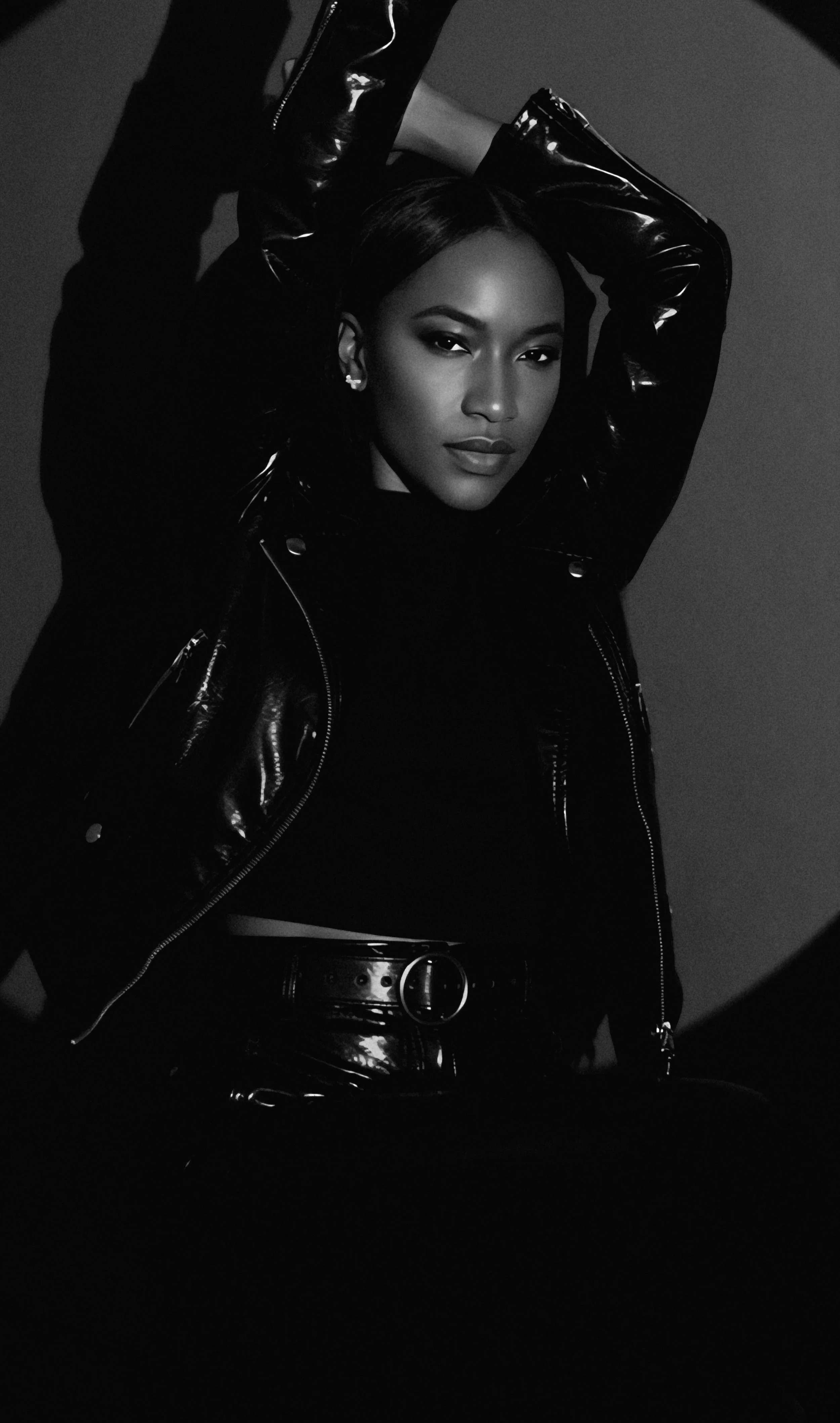 Black and white photo of a woman with short hair, wearing a shiny leather jacket, with her arms raised above her head.