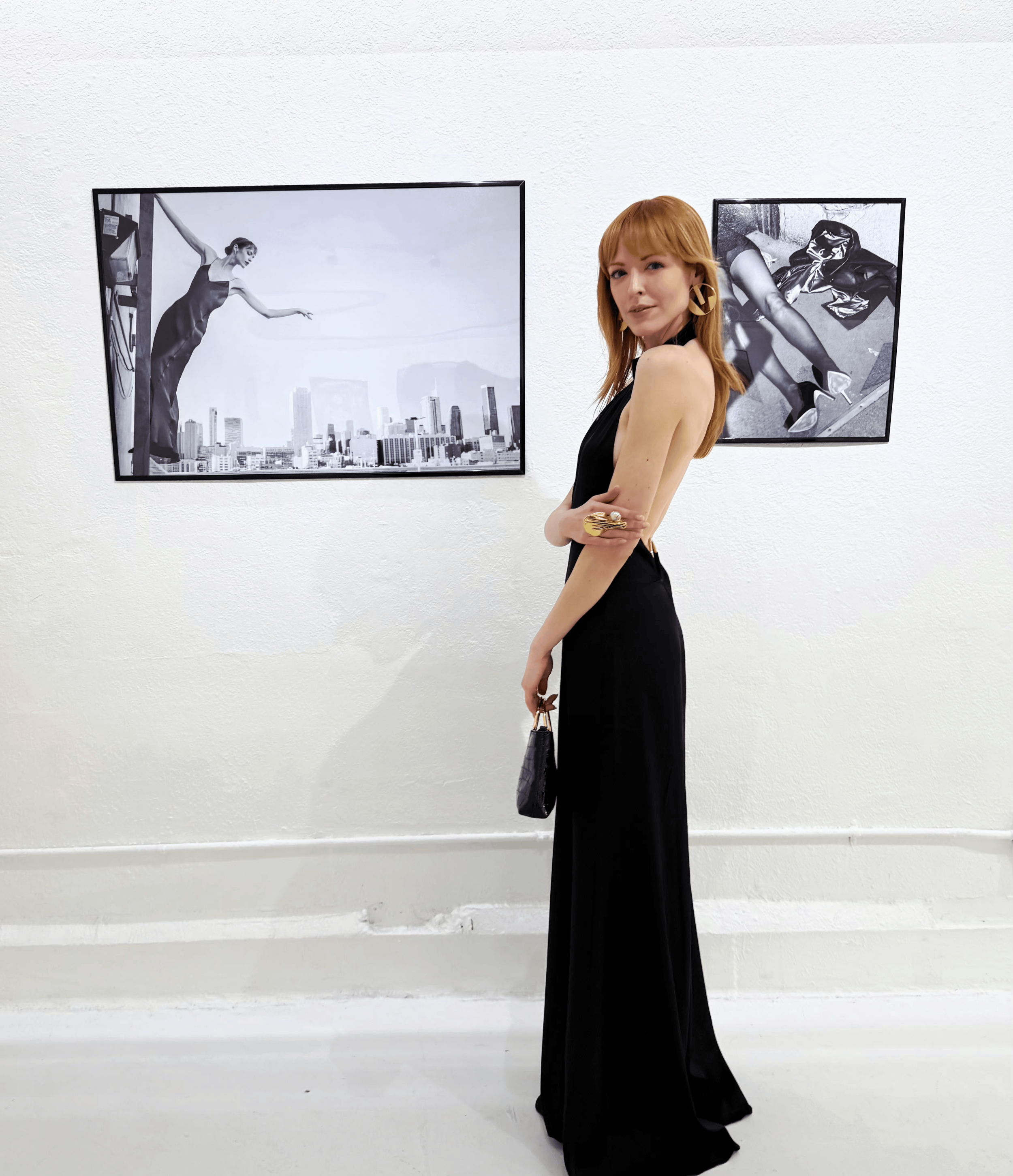 A woman with red hair in a black dress stands in an art gallery, looking at black-and-white photographs on the wall.