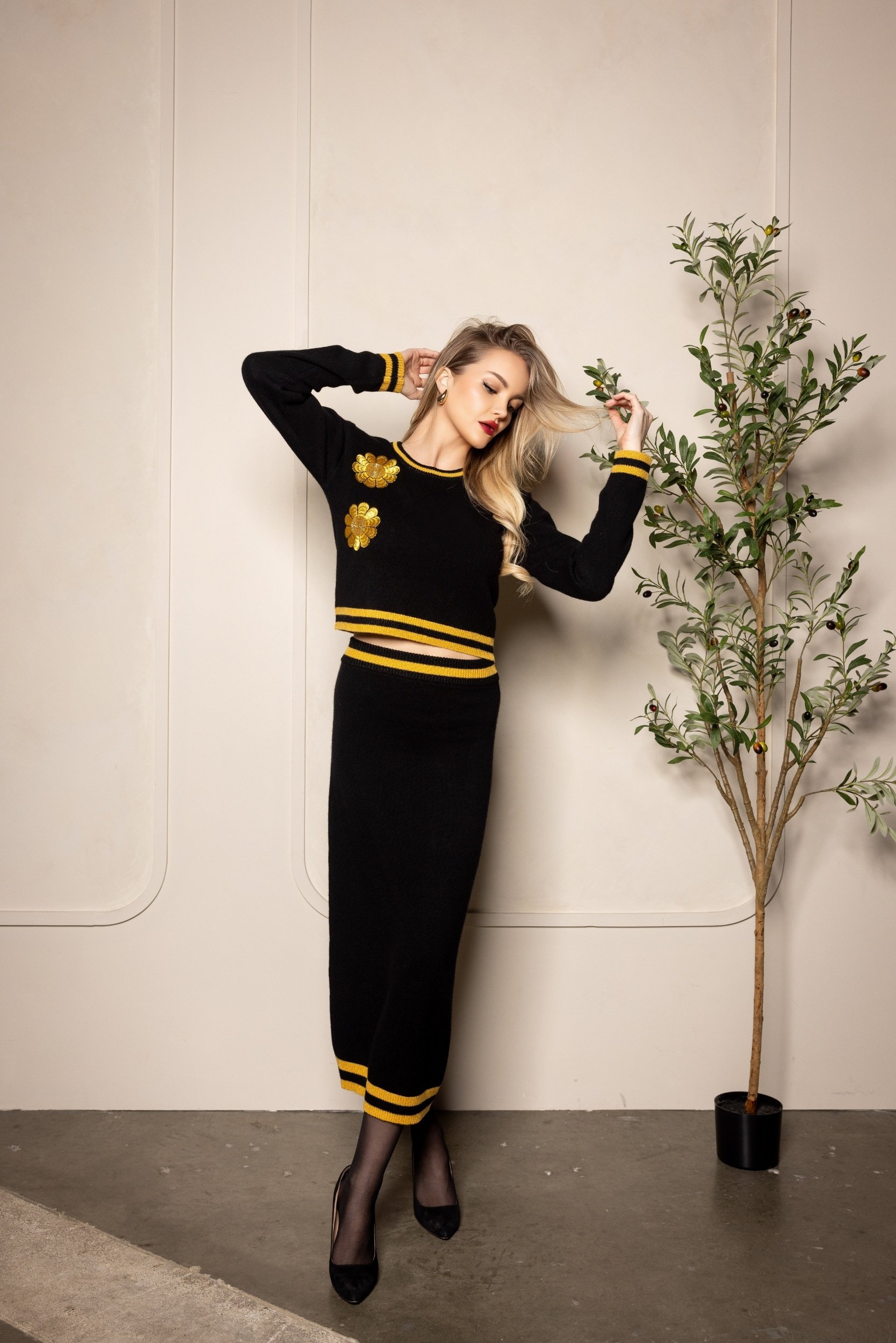 blonde woman wearing black ritzy cashmere outfit with long black pencil skirt accented with gold with hand on plant looking to the right in white background.