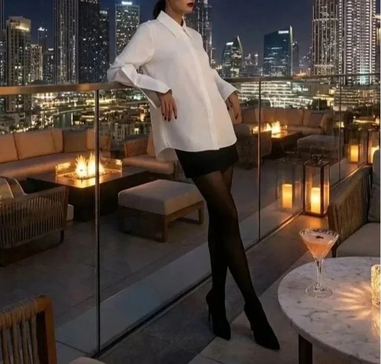 Woman in a white blazer, black shorts, and black sheer tights posing on a rooftop at night with city skyline in the background, surrounded by dimly lit candlelit furniture and a martini glass on a marble table.