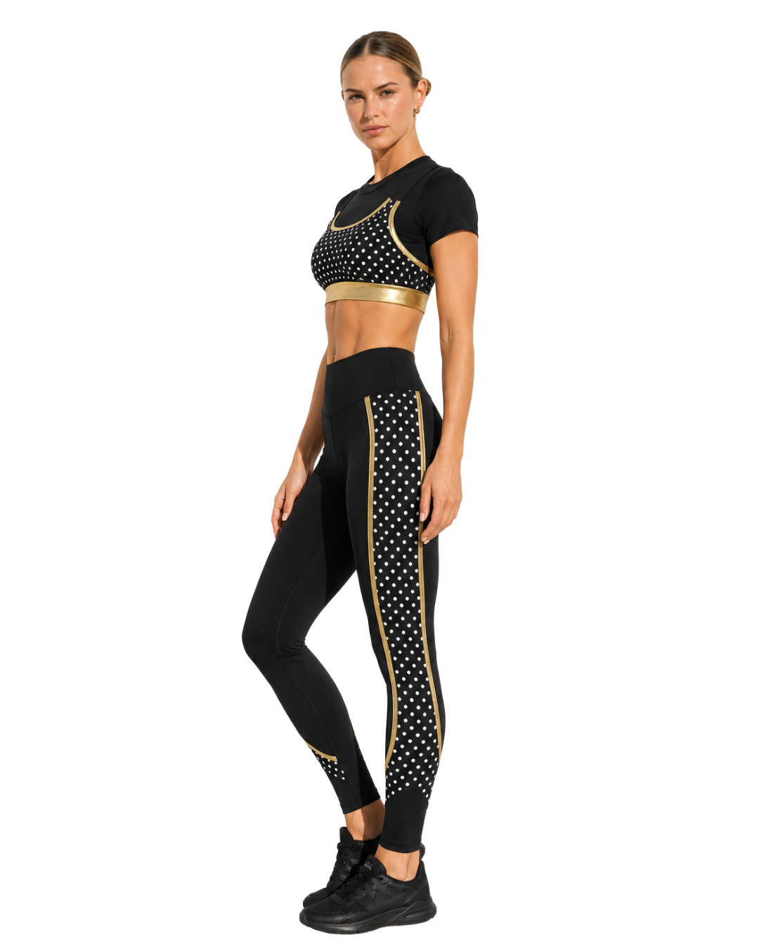 A woman in black athletic wear with gold accents and polka dot patterns, standing with one hand on her hip, against a plain white background.