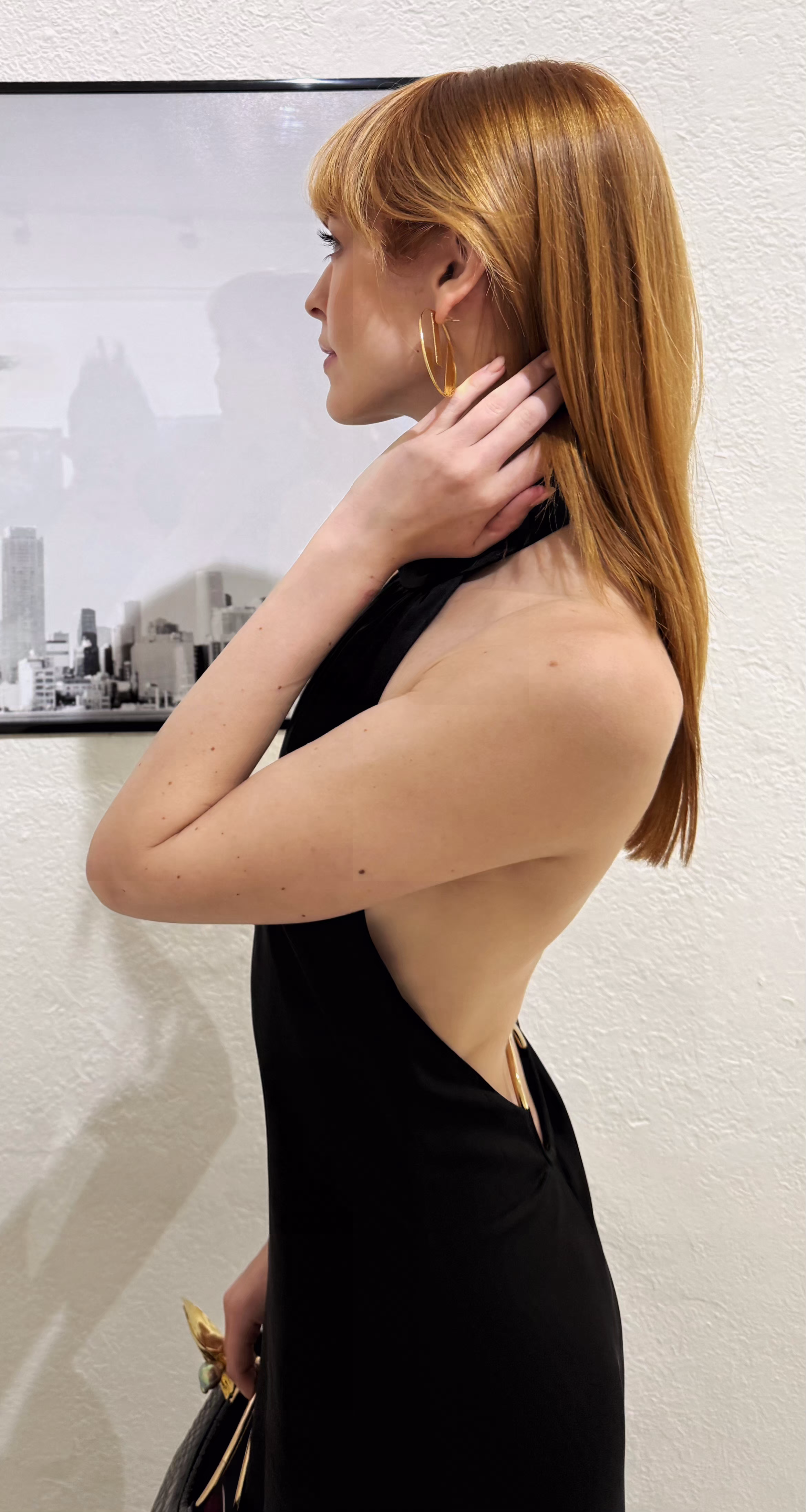 A woman with red hair and gold hoop earrings is standing sideways against a white wall, touching her neck with her right hand, wearing a sleeveless black dress with a revealing side cutout.