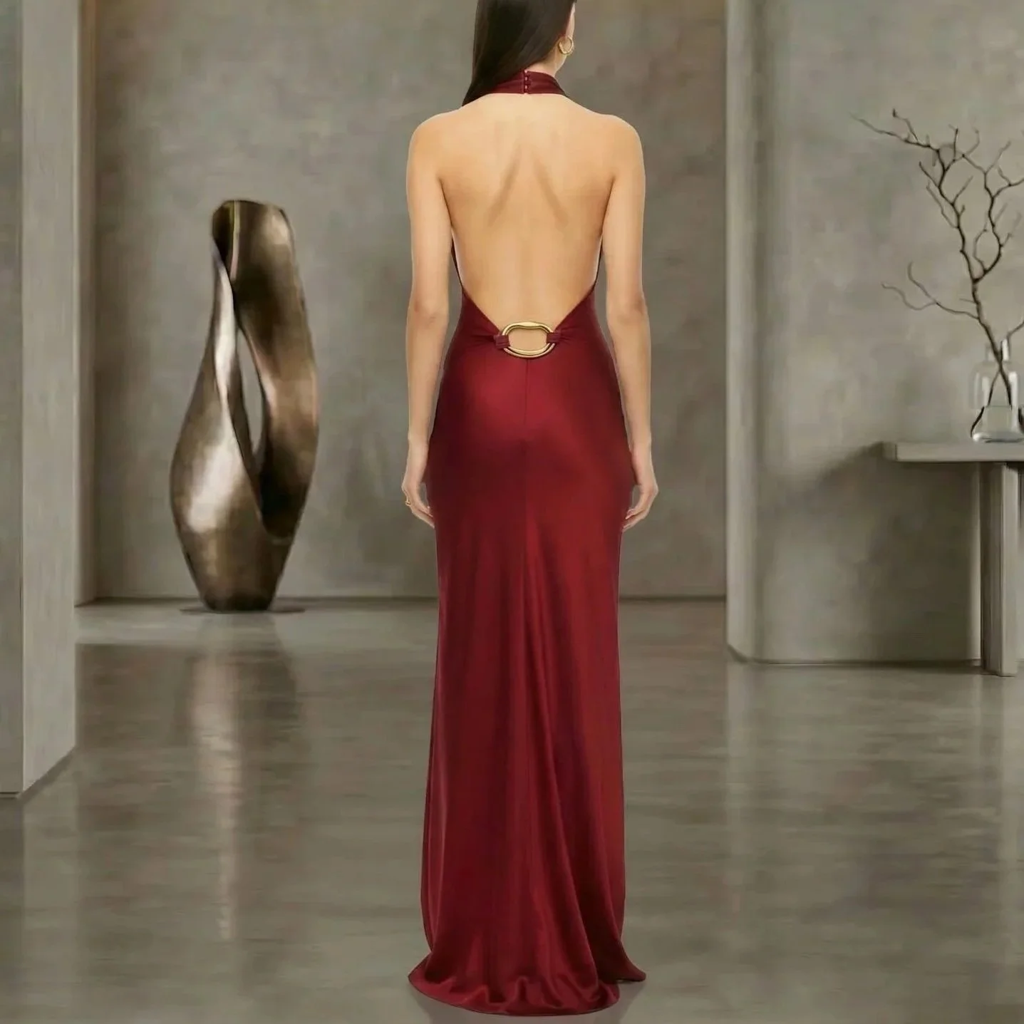 Back view of a woman in a long red gown with an open back and a gold ring detail at the waist, standing in a modern room with minimalist decor.