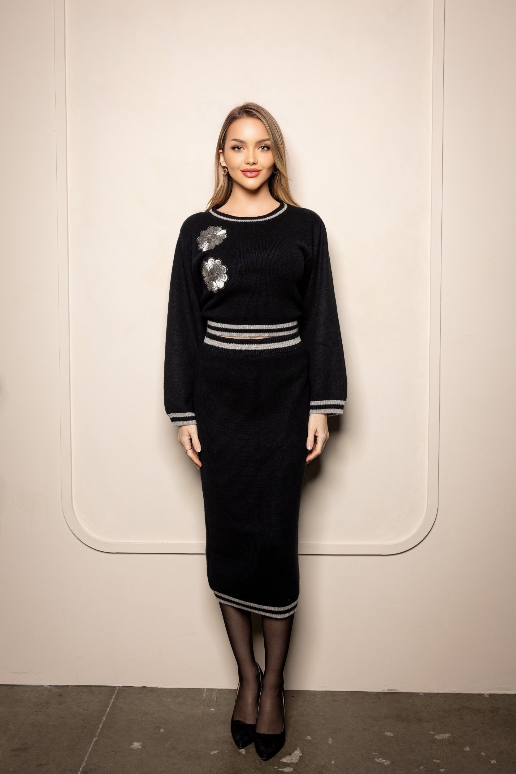 blonde woman wearing black ritzy cashmere outfit with long black pencil skirt accented with silver looking at camera in white background.