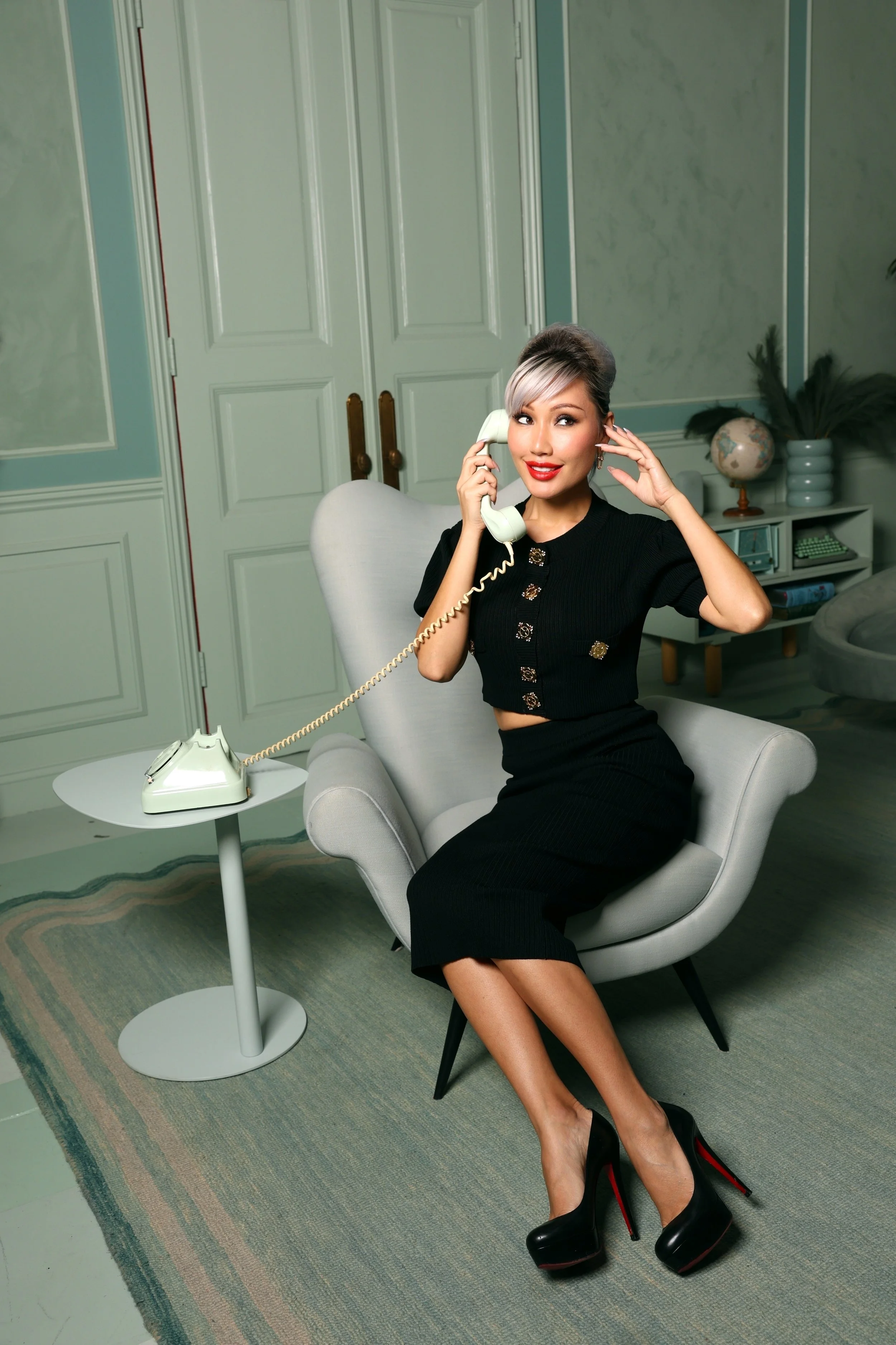A woman with short, ombre hair, wearing a black two-piece outfit and black high heels, sitting in a modern living room while talking on a vintage rotary phone.