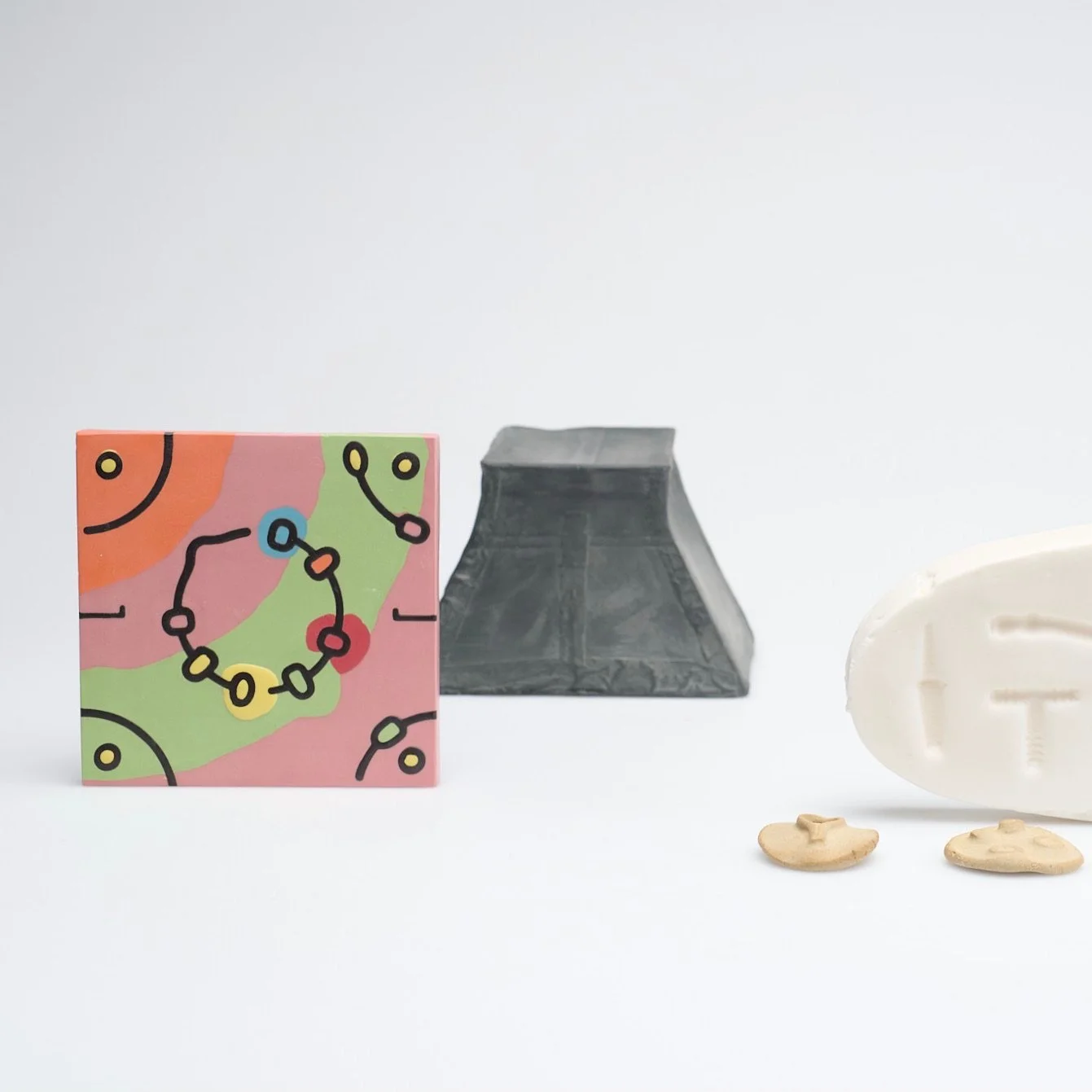Decorative objects including a small colorful box with abstract designs, a black rectangular block, two oval white soap bars with raised geometric patterns, a beige oval soap bar with similar patterns, and two small beige soap chips on a white backgr