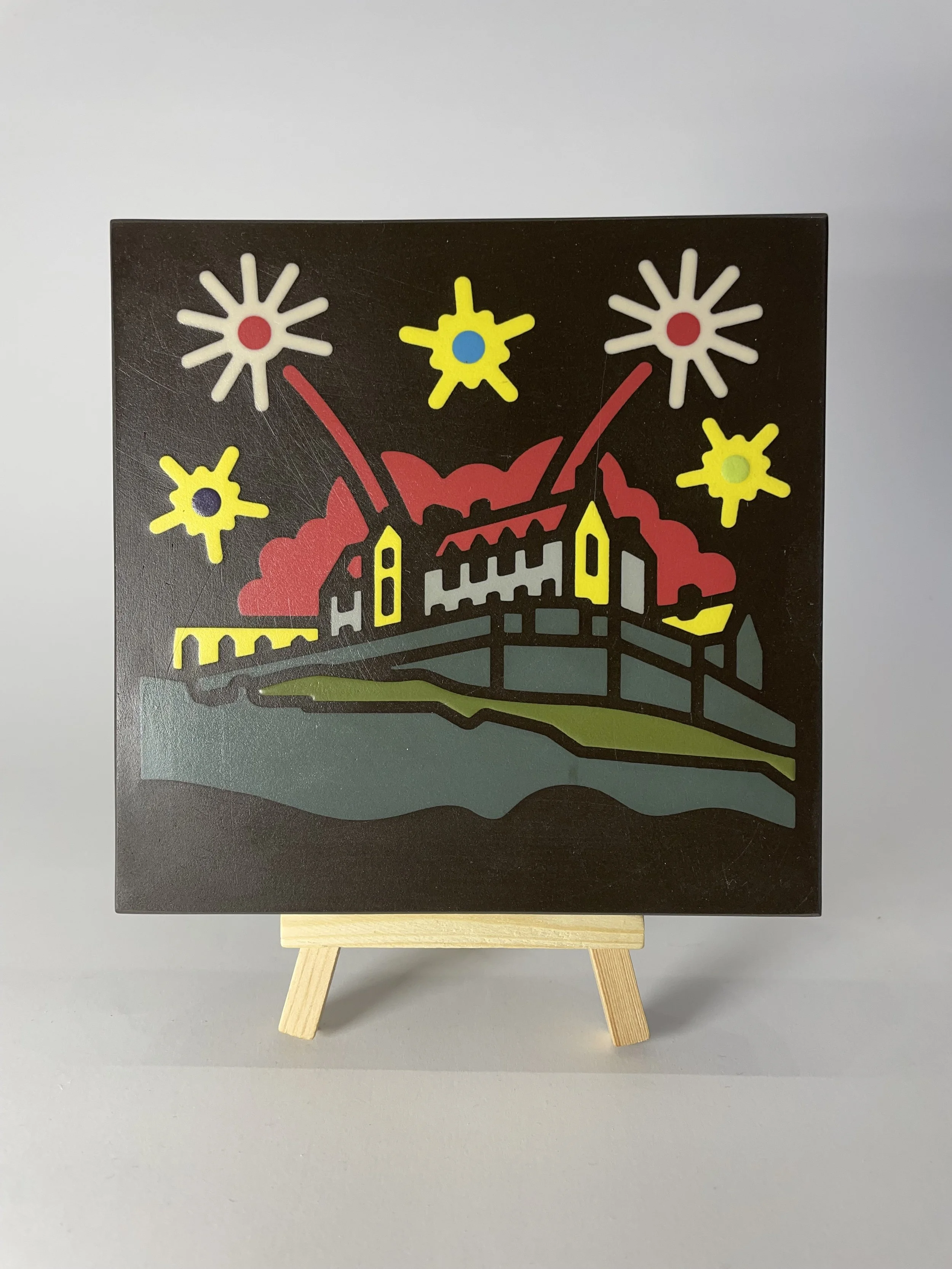Colorful encaustic tile depicting a stylized castle on a hill with fireworks or explosions above it, set against a black background. Mounted on a small wooden easel.