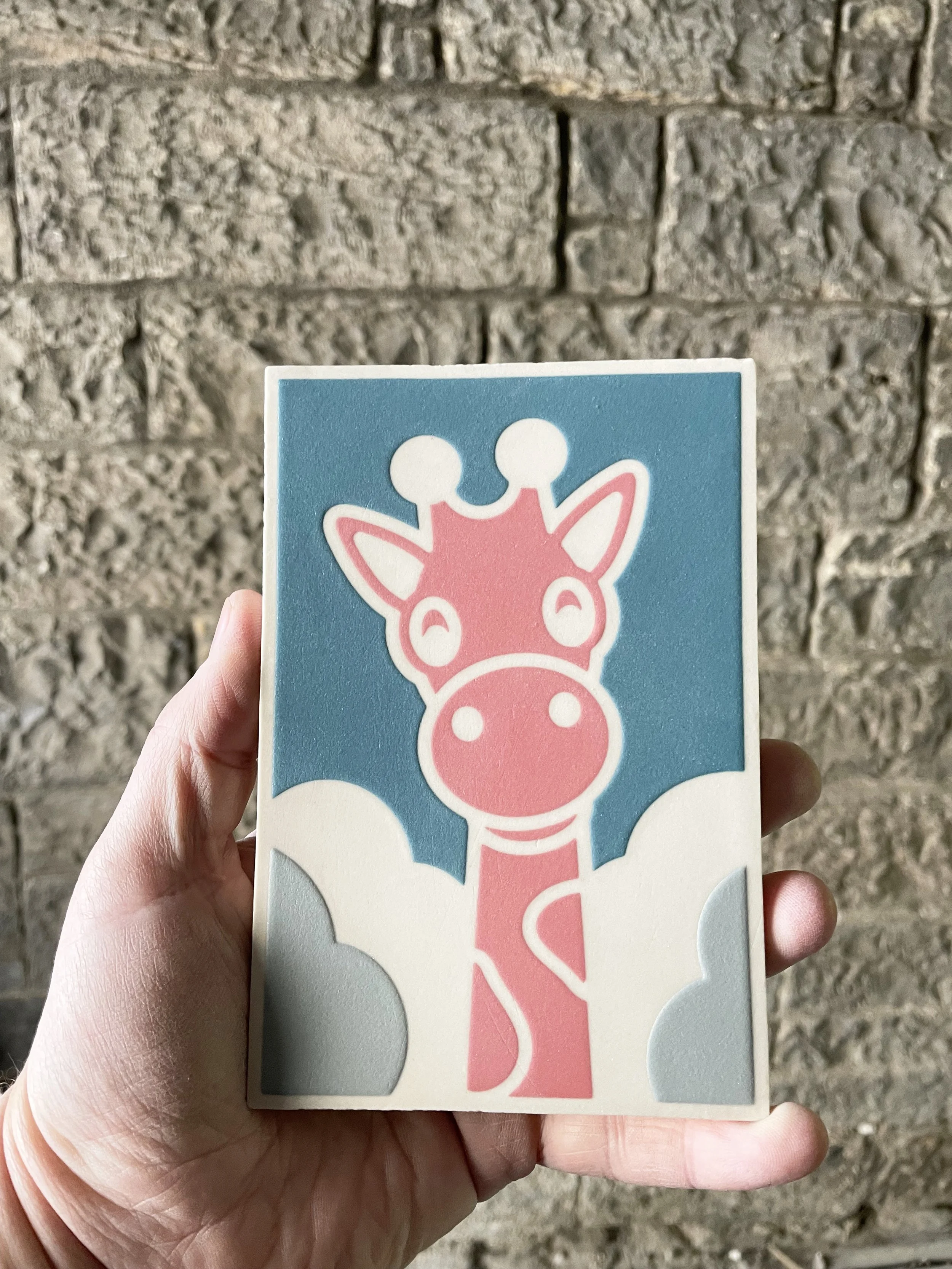 Person's hand holding a white, blue, pink and grey encaustic tile depicting a cartoon giraffe in the clouds. Sandstone wall background.