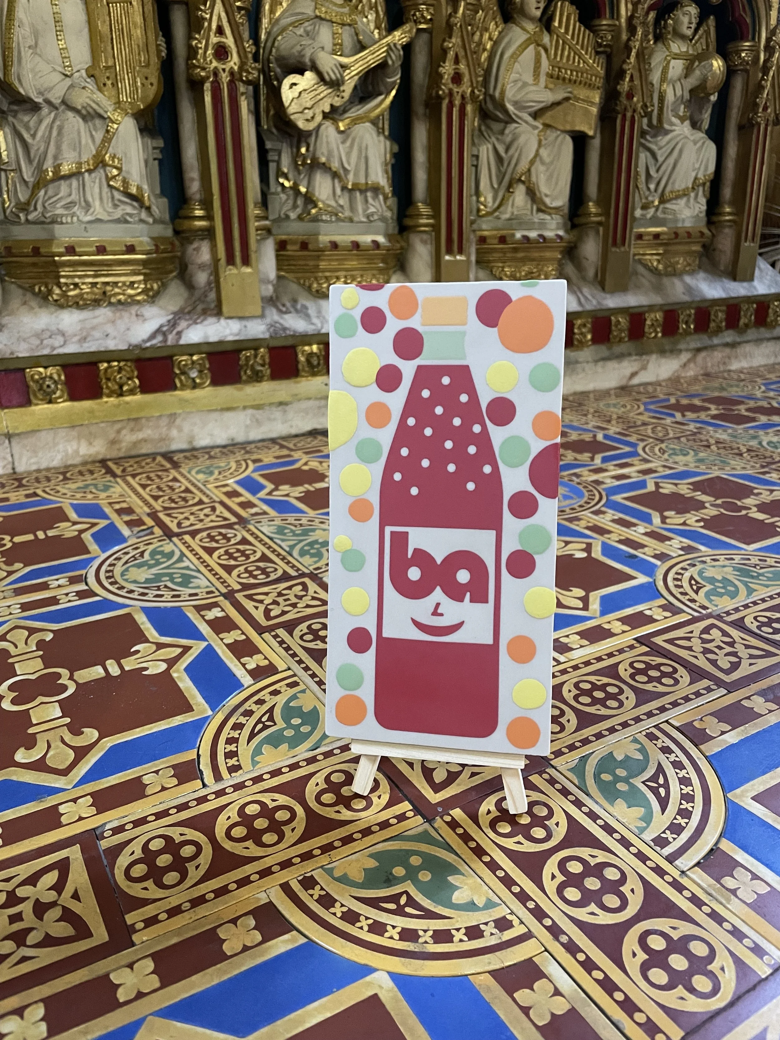 Colorful encaustic tile of a smiling red fizzy drink bottle with bubbles on it, surrounded by multicolored circles, on a stand inside a room with ornate, gold-trimmed sculptures and decorative tiled floor.