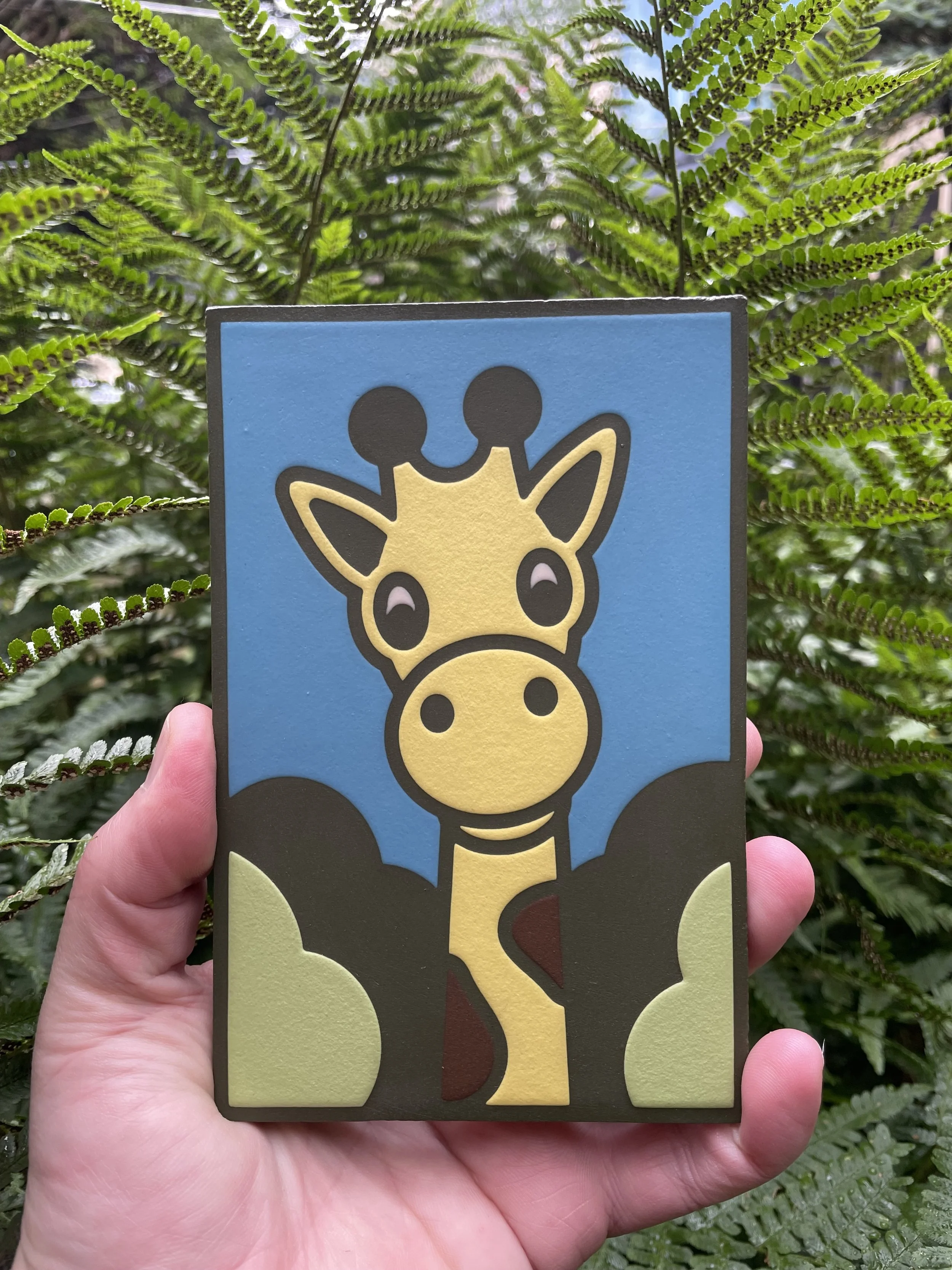 Encaustic tile with a cartoon-like illustration of a giraffe with a blue sky background, green bushes, and dark trees.