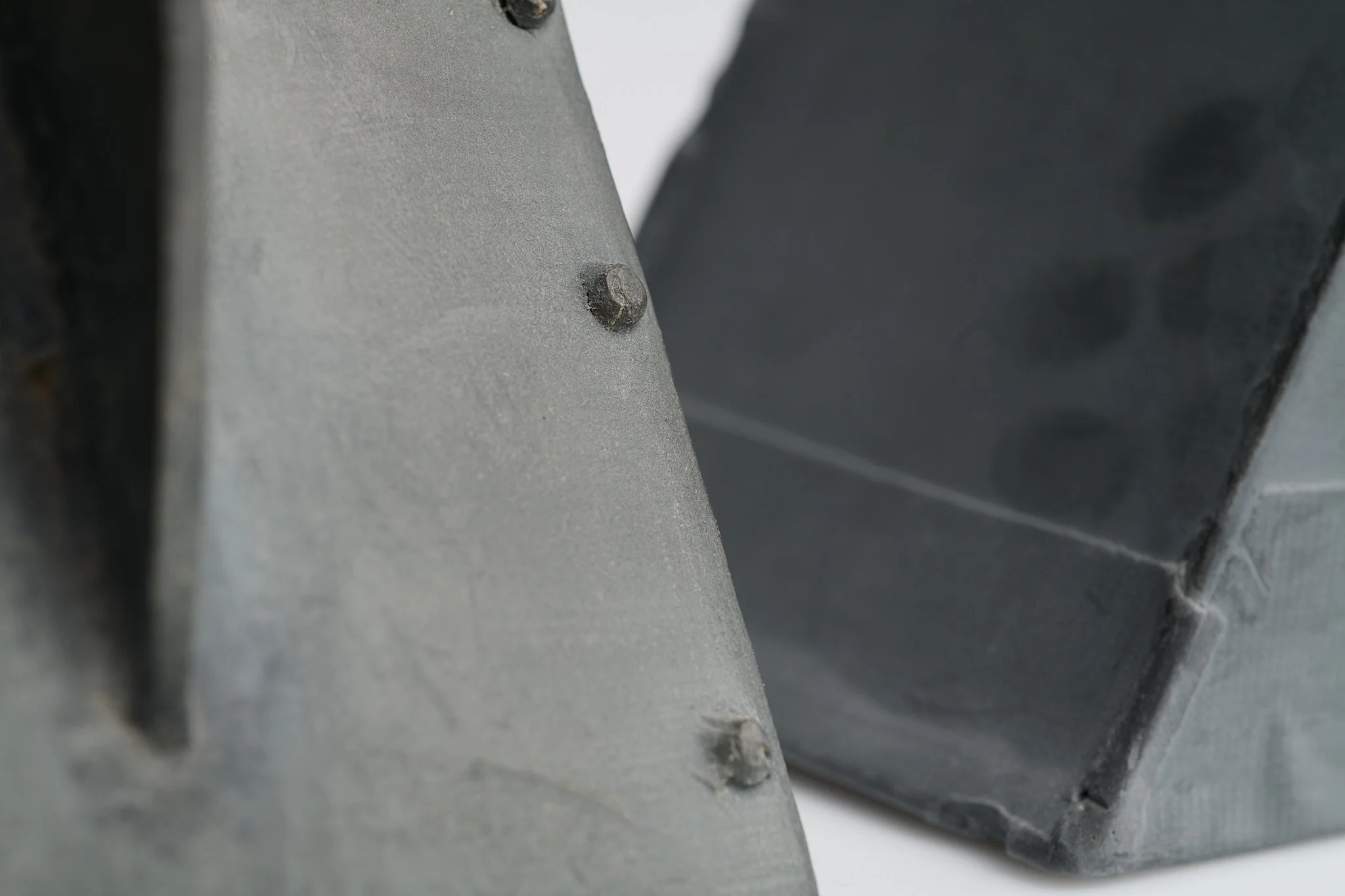 Close-up of two metallic looking ceramic objects with visible rivets and textured surfaces, one gray and the other darker gray.