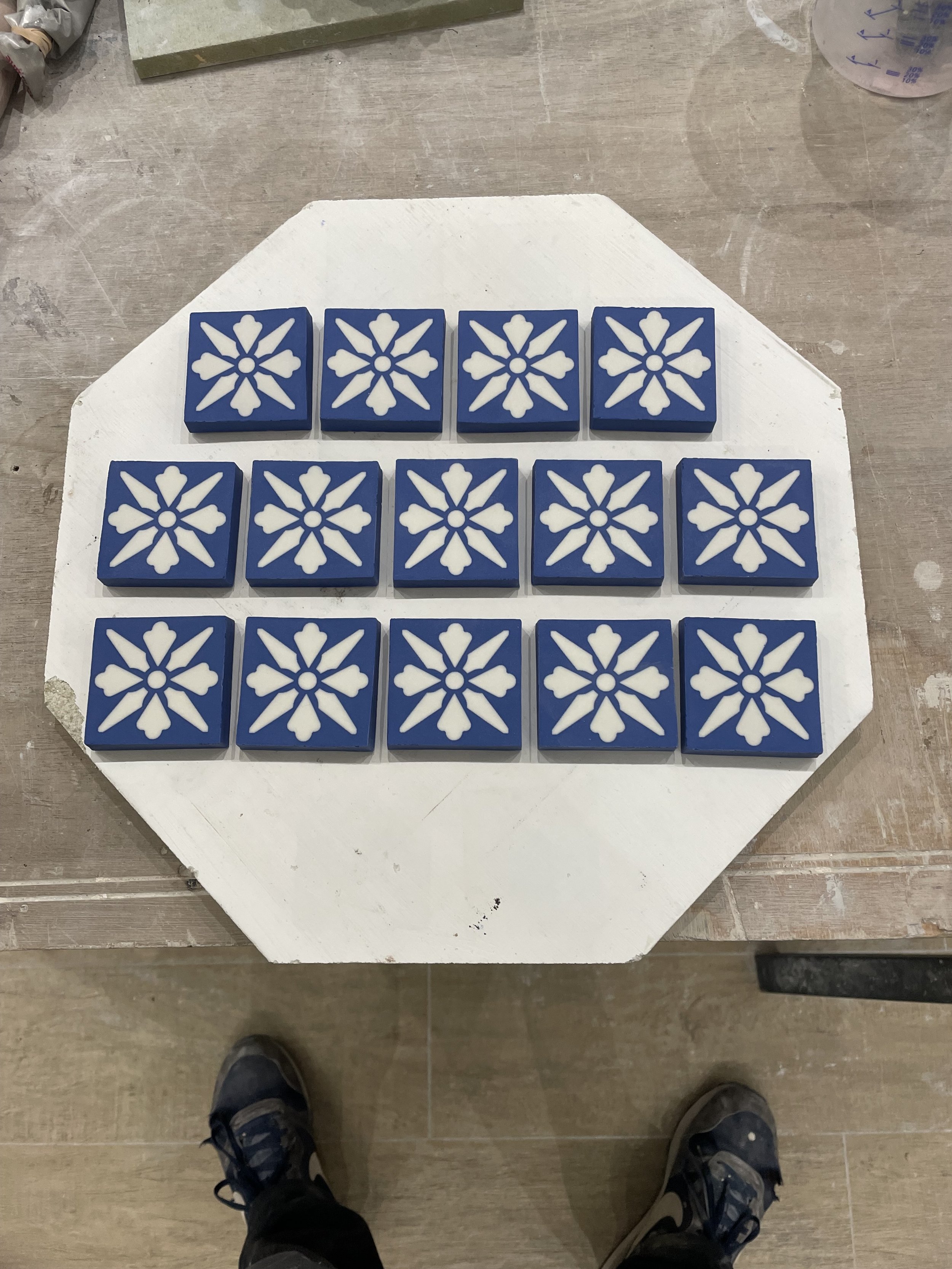 A hexagonal white kiln shelf with a 3x6 grid of blue tiles featuring white snowflake patterns. The person's shoes are visible at the bottom of the image.