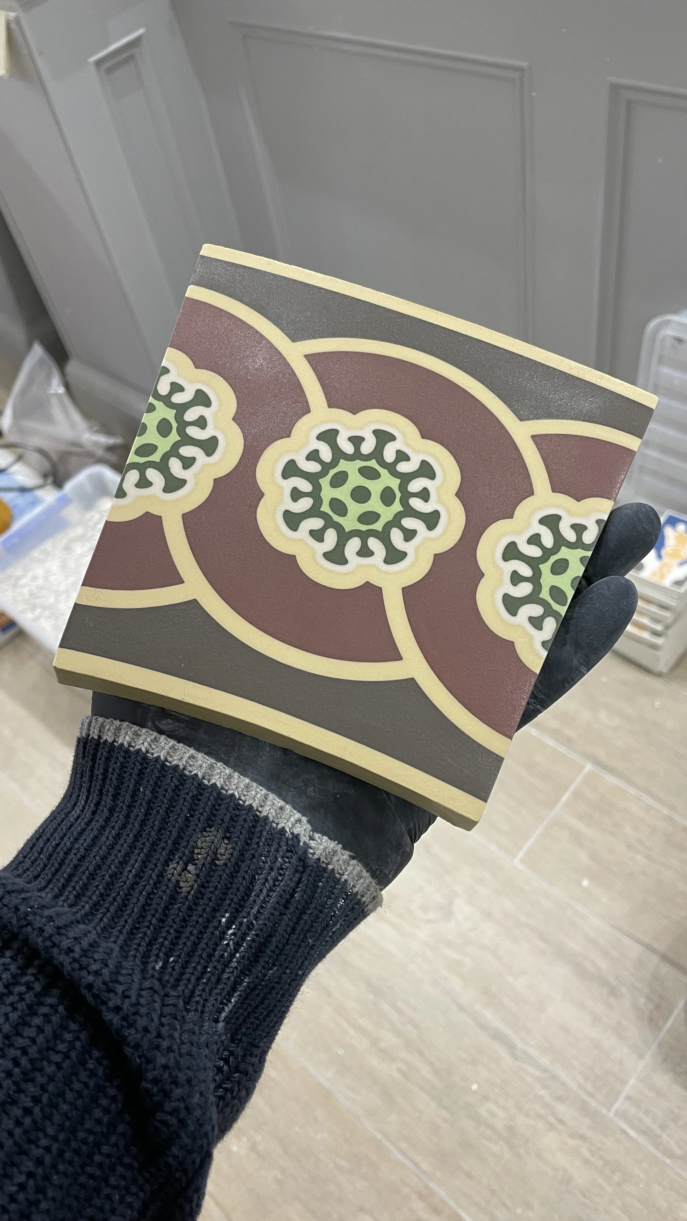 A person wearing a black glove is holding a small, square tile with a decorative pattern in shades of green, black, red, and yellow, featuring stylized floral designs.