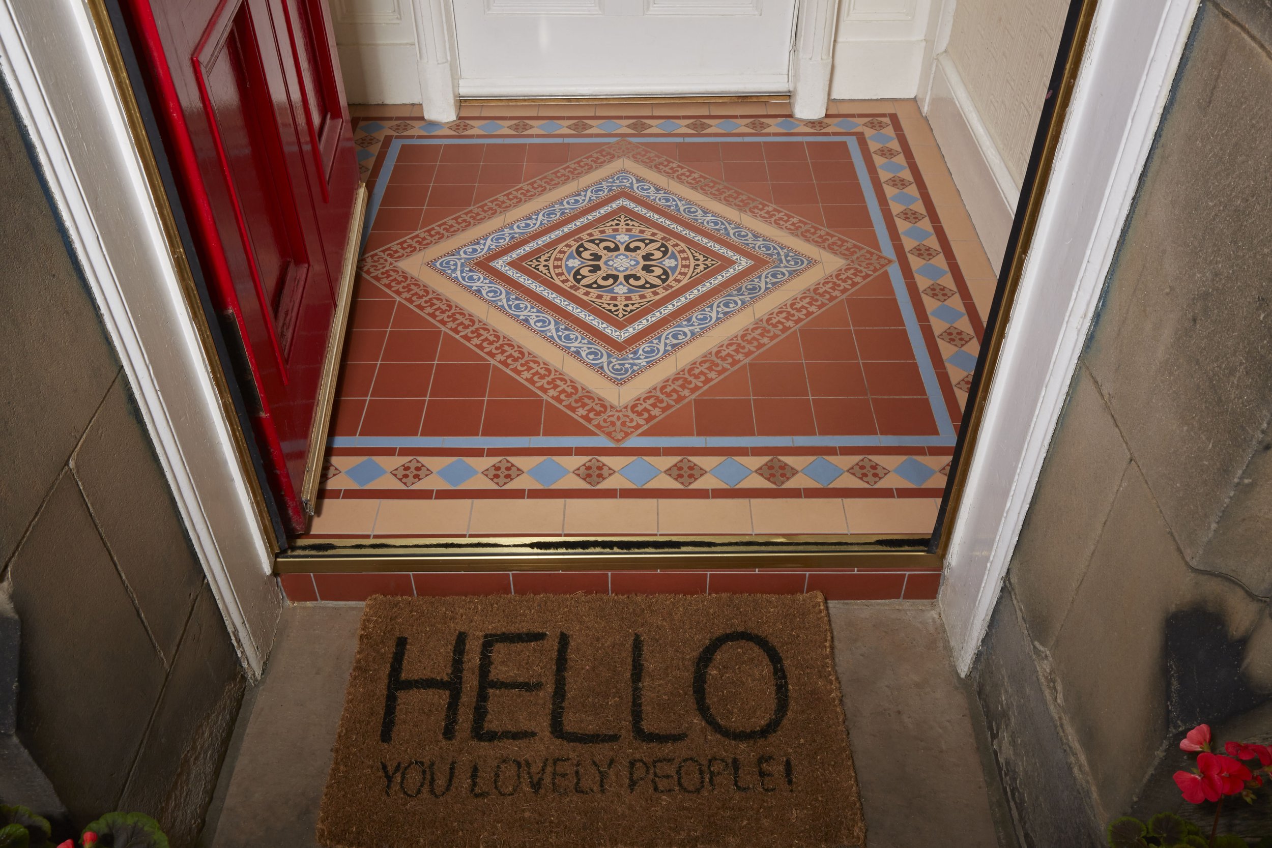View of a decorative tiled floor at the entrance of a house with a brown doormat that says 'HELLO YOU LOVELY PEOPLE!' in front of a red door. The floor has intricate geometric and floral tiles in shades of red, cream, blue, and black.