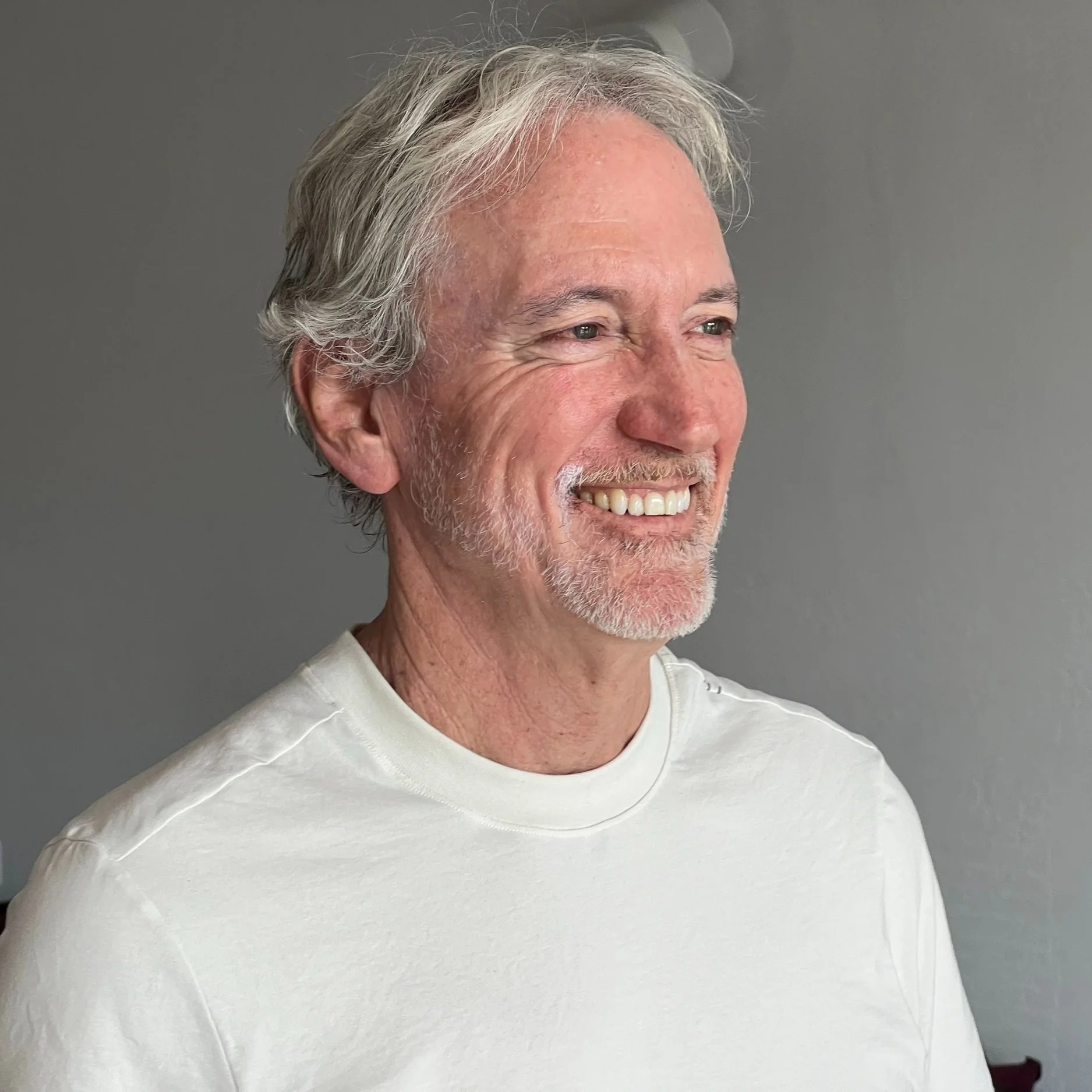 Smiling elderly man with gray hair and beard, wearing a white shirt, standing against a gray wall.