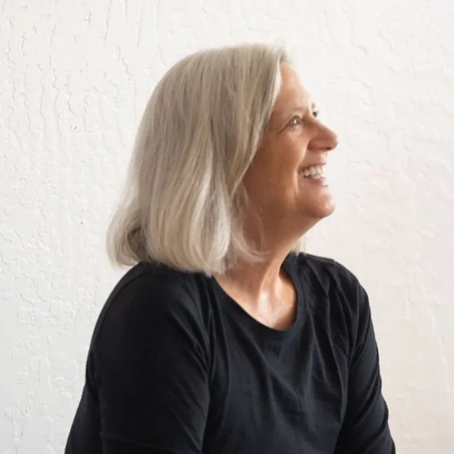 Profile of a middle-aged woman with shoulder-length gray hair, smiling and looking to the side, wearing a black shirt against a white textured wall.