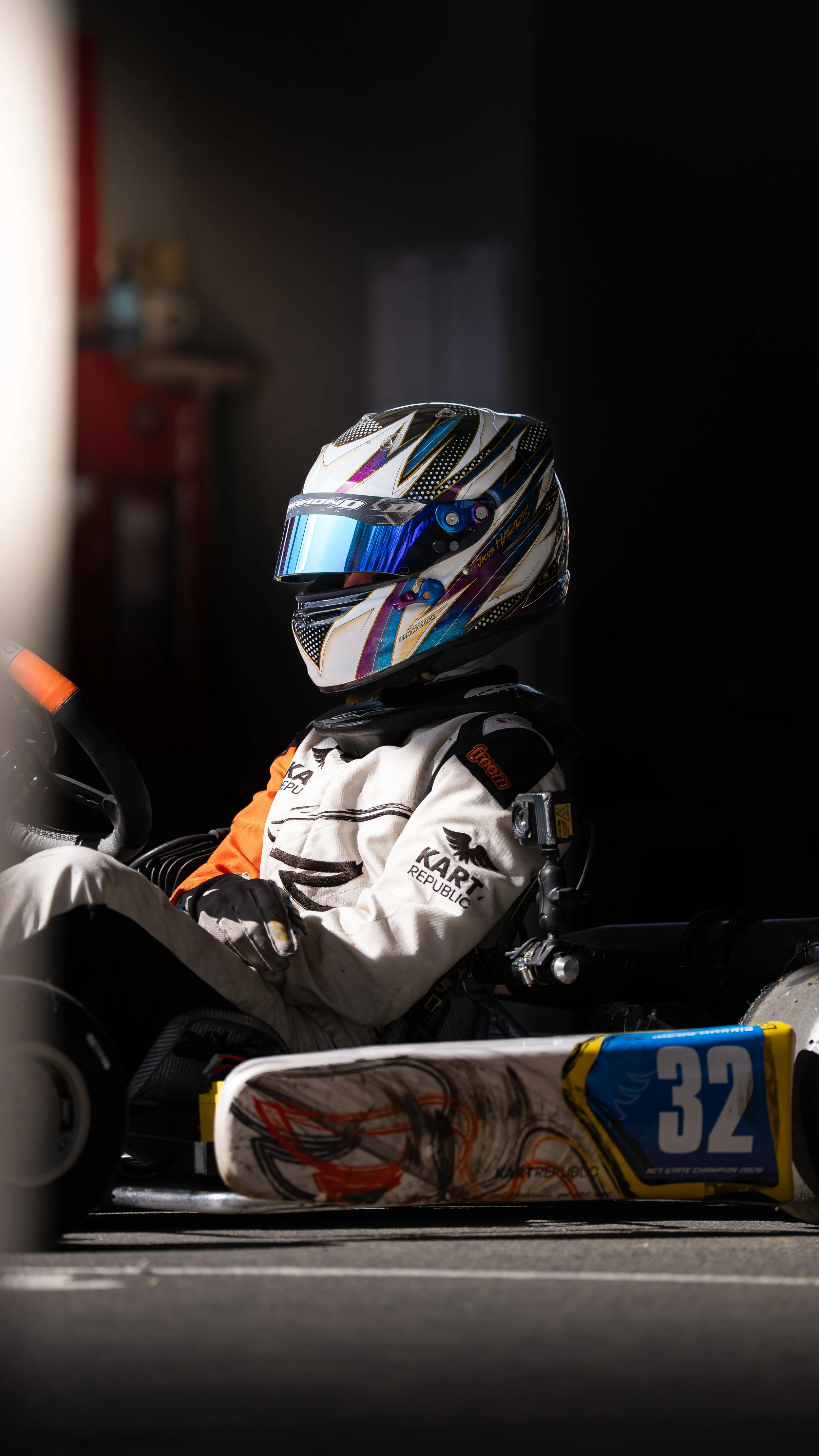 A race car driver wearing a colorful helmet and racing suit sitting in a race car, with parts of the track and surrounding environment blurred in the background.
