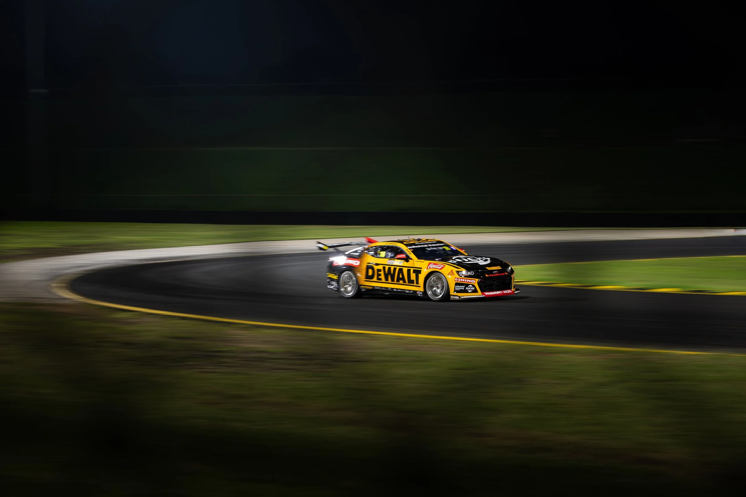 A yellow race car with sponsorship decals speeds around a dark race track at night, with motion blur indicating high speed.