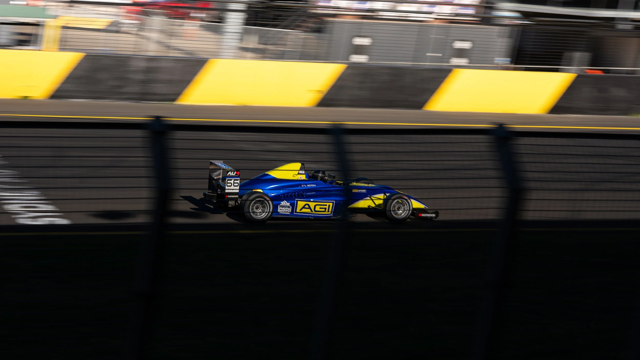 A blue and yellow race car speeding on a racetrack, blurred background, with barriers and fencing visible.