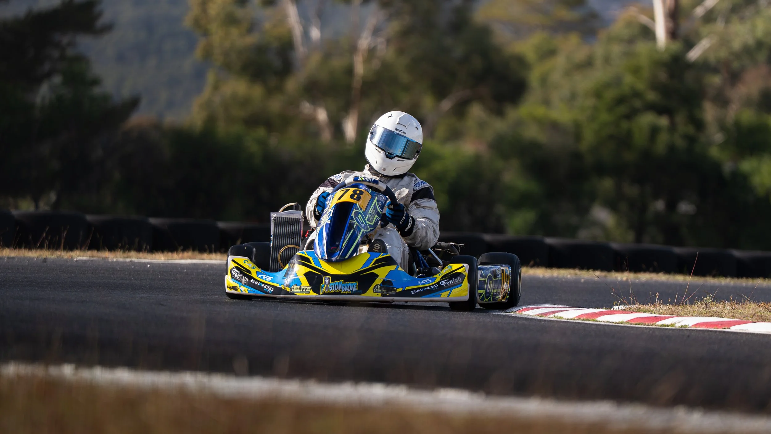 A person wearing a white helmet and racing gear driving a yellow go-kart on a race track.