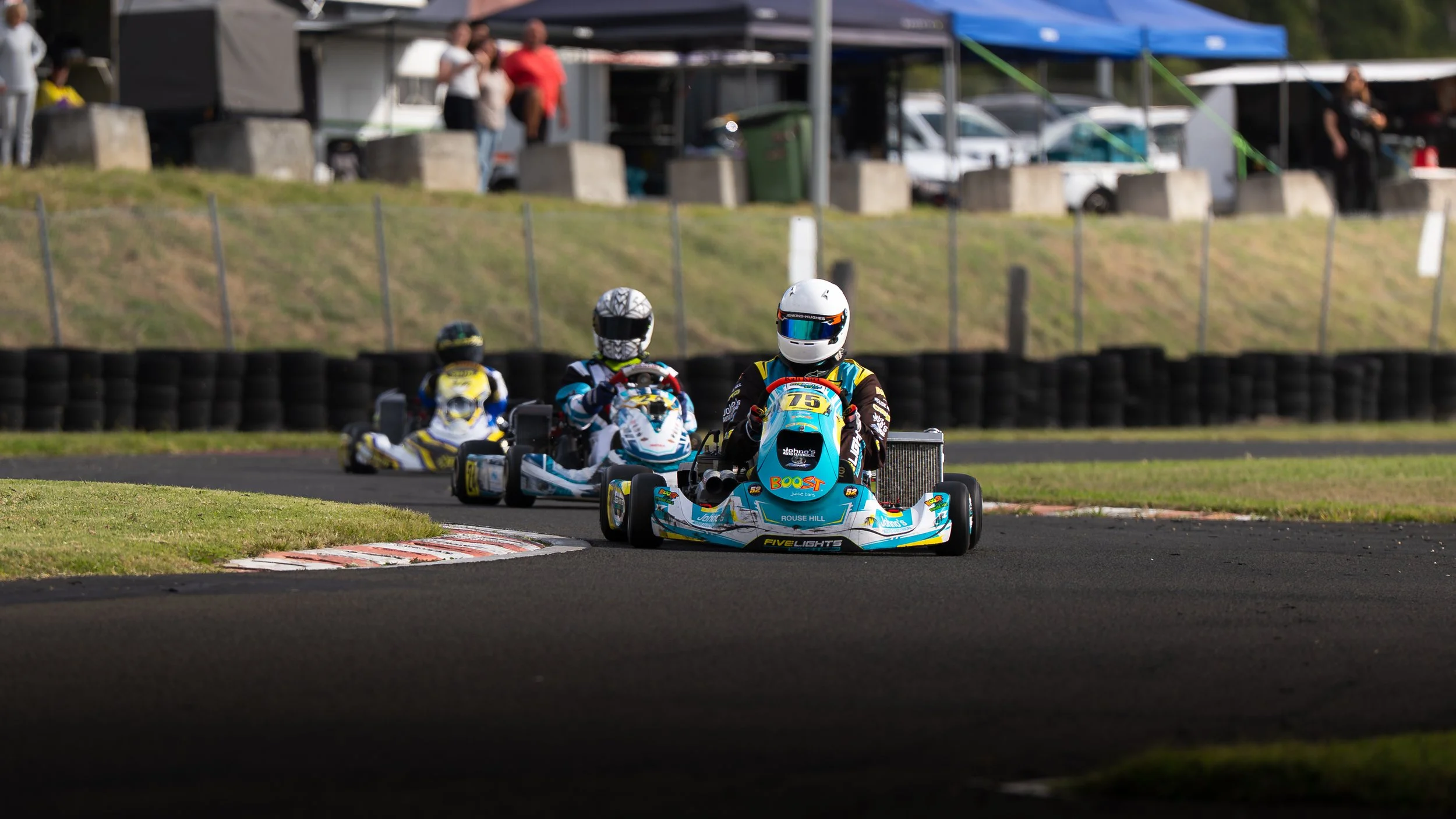 Three go-karts racing on an outdoor track with drivers wearing helmets and racing suits, with spectators and tents in the background.