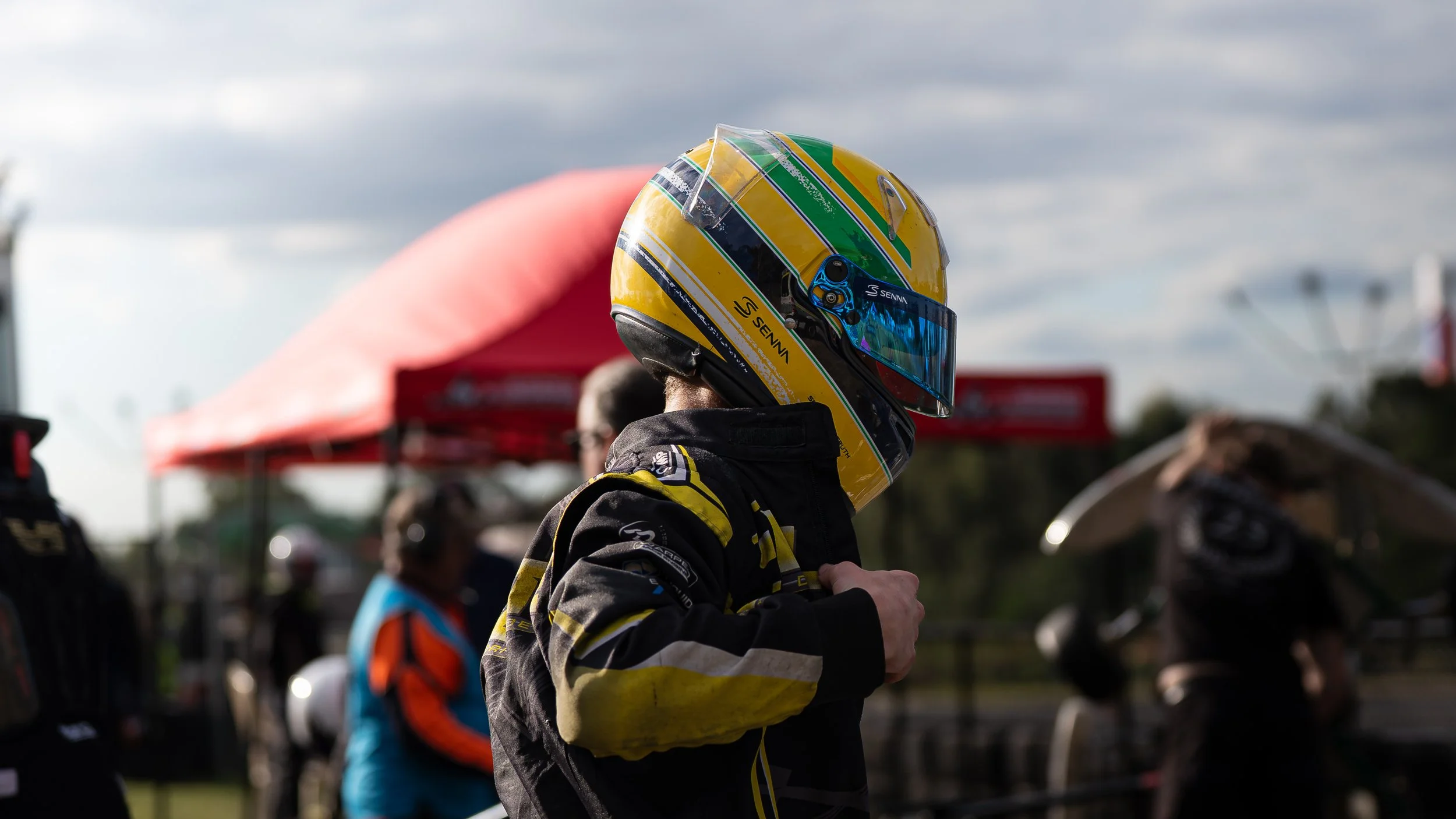 Person wearing a racing helmet and racing suit, standing outdoors at a racing event.