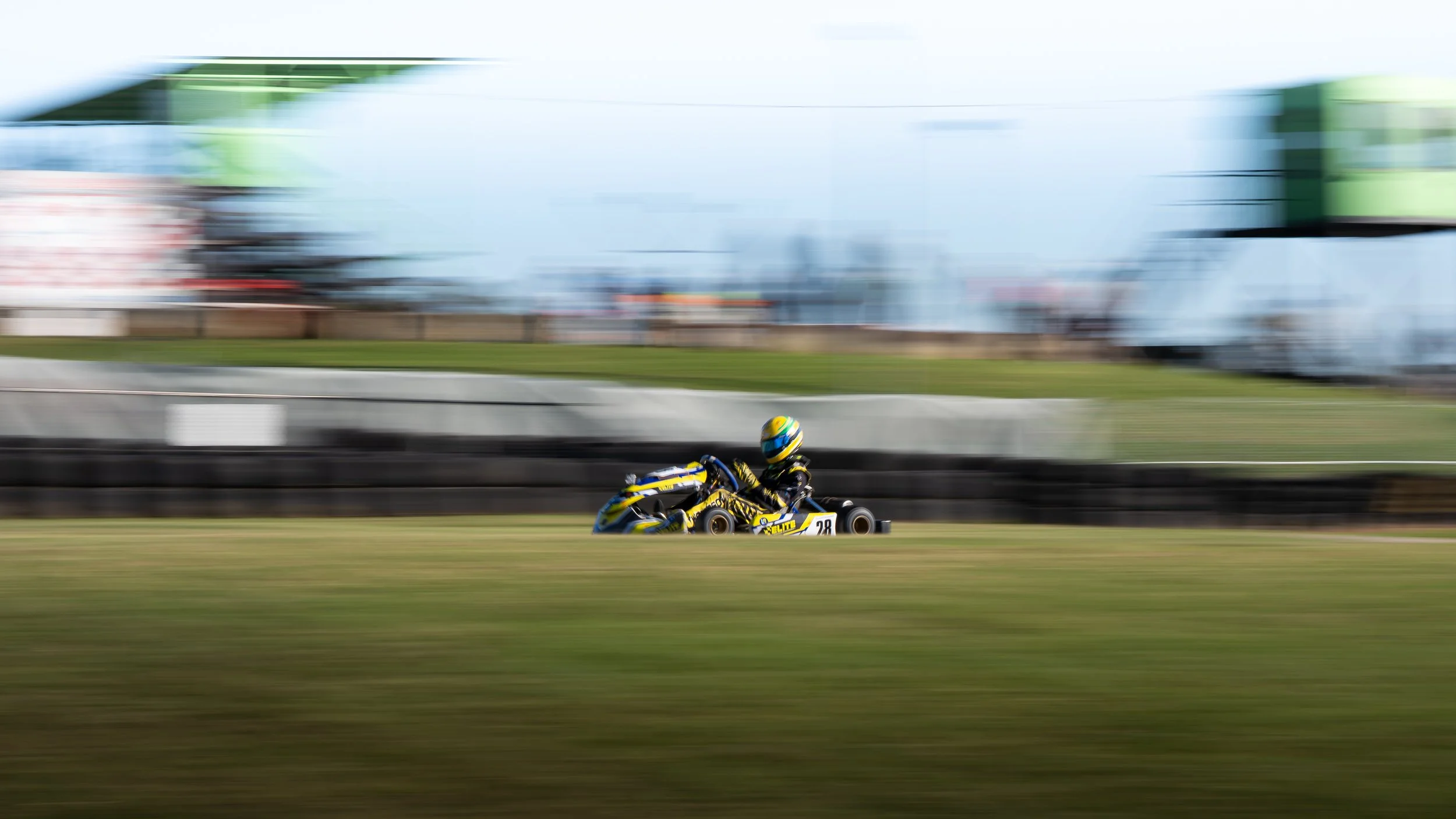 A go-kart racer wearing a colorful helmet and a racing suit speeds along a track, creating a motion blur effect in the background.