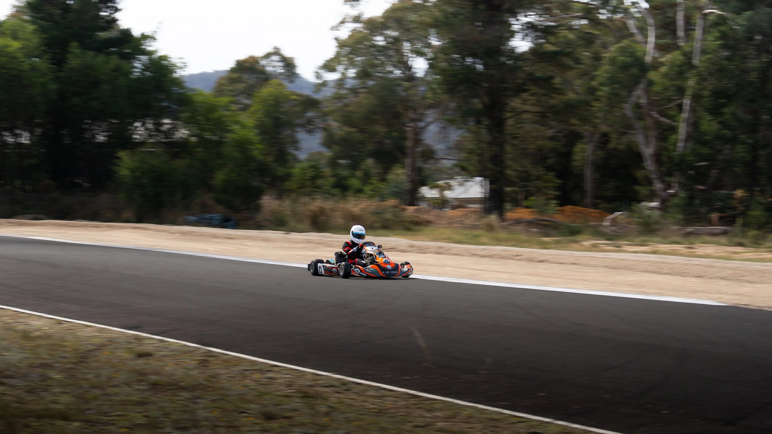 A person in a helmet driving a go-kart on a race track with trees in the background.