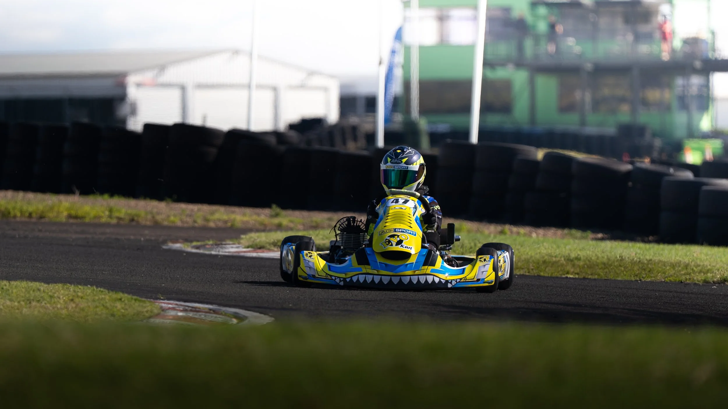 A go-kart with a yellow and blue design, featuring a jagged mouth graphic, on a race track with a driver wearing a helmet. The background shows tire barriers and industrial structures.