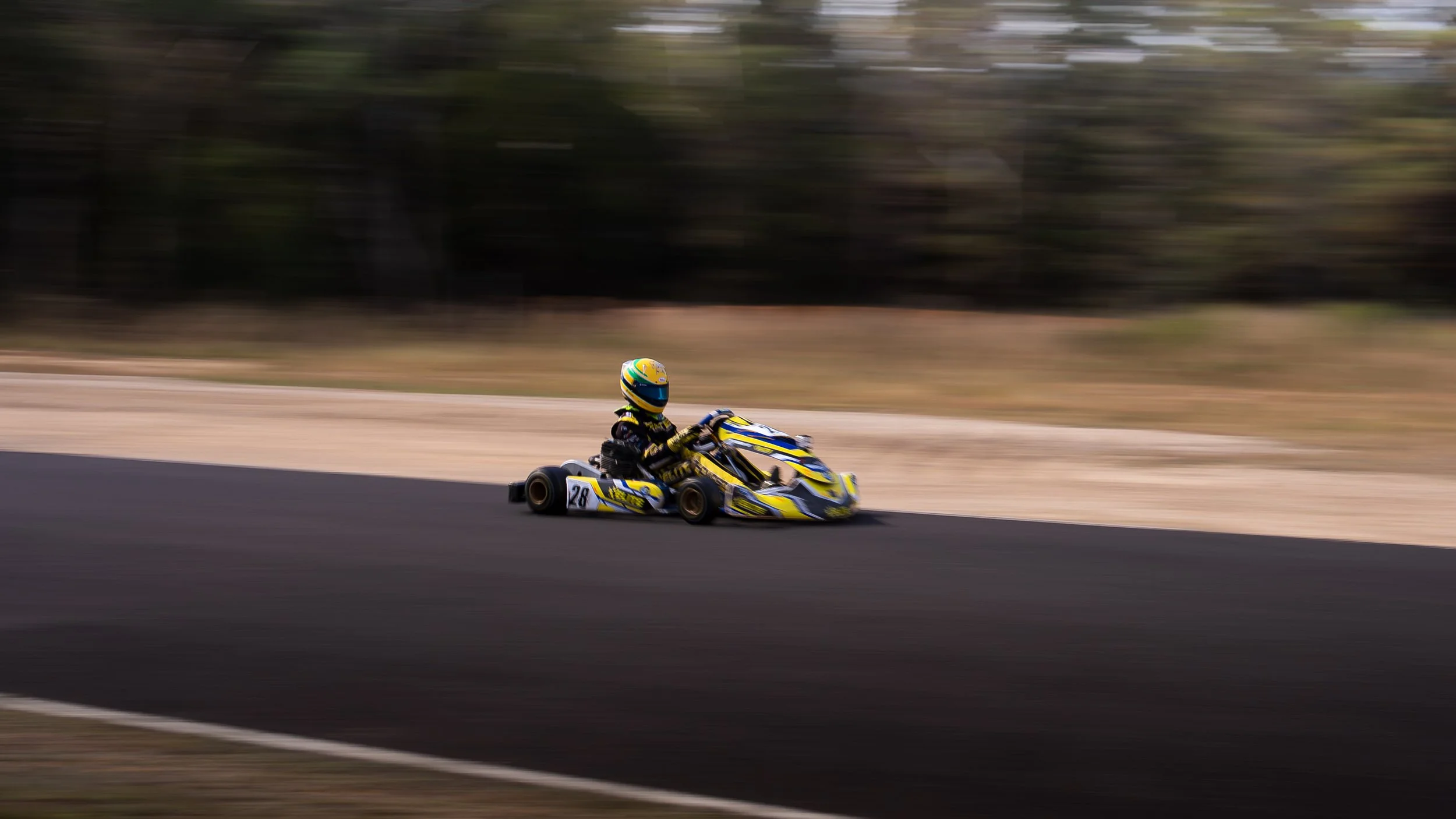 A go-kart racer wearing a yellow and blue racing suit and helmet, speeding on a racetrack with a blurred background.