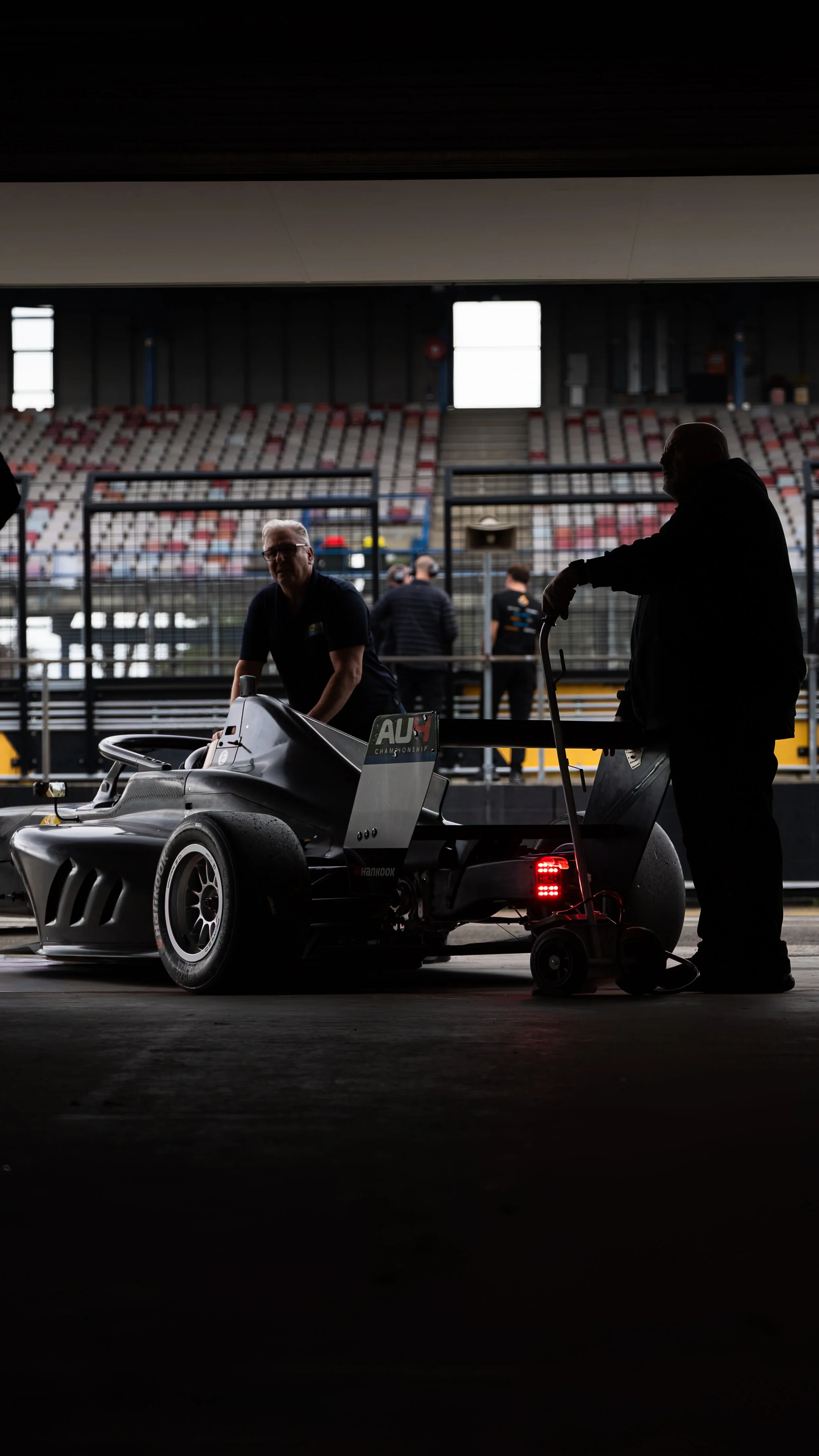 Two men working on a race car inside a garage at a racing track, with empty grandstands in the background.