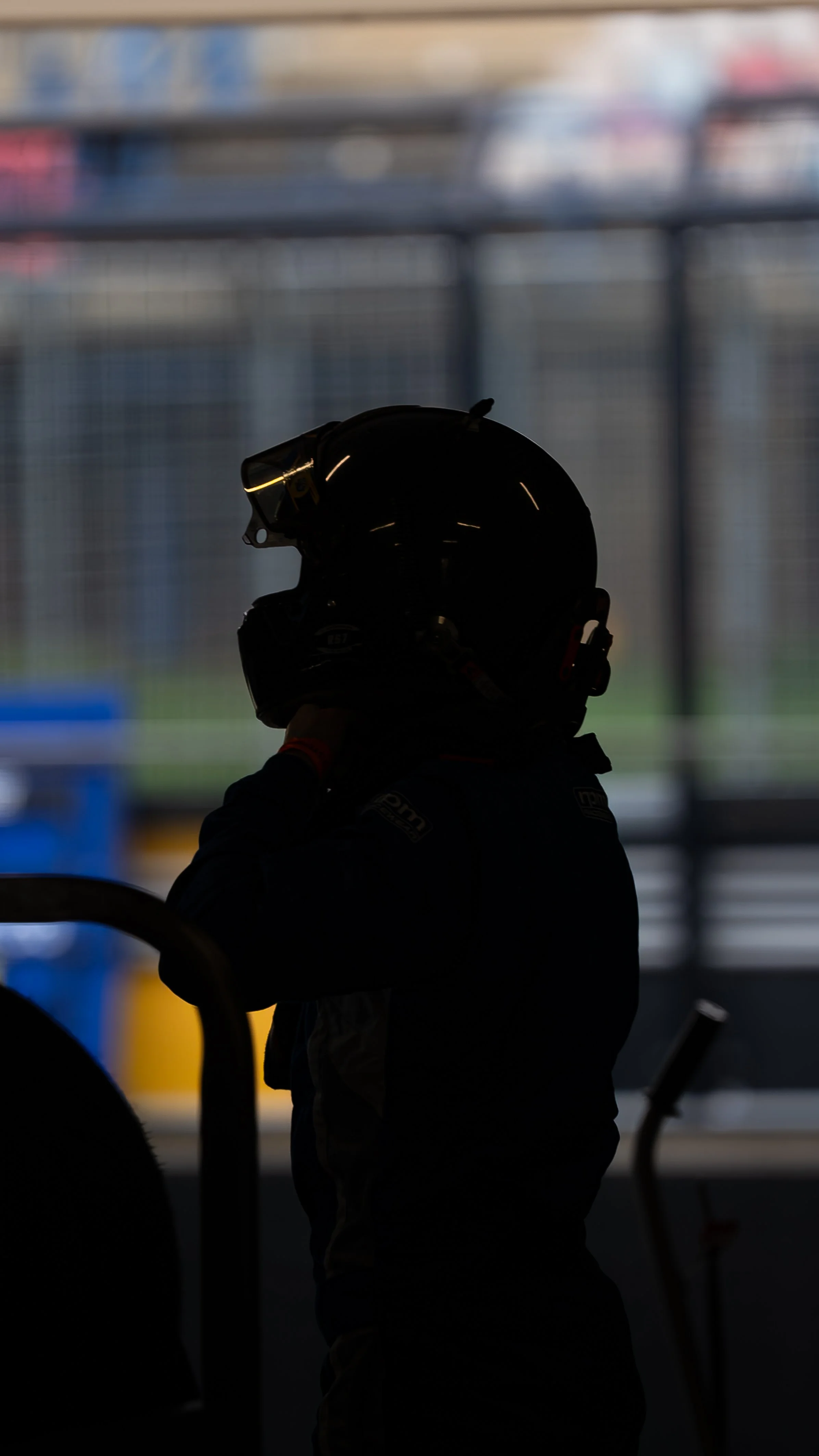 Silhouette of a person wearing a racing helmet, standing in a dimly lit area with a blurred background of shelves or structures.