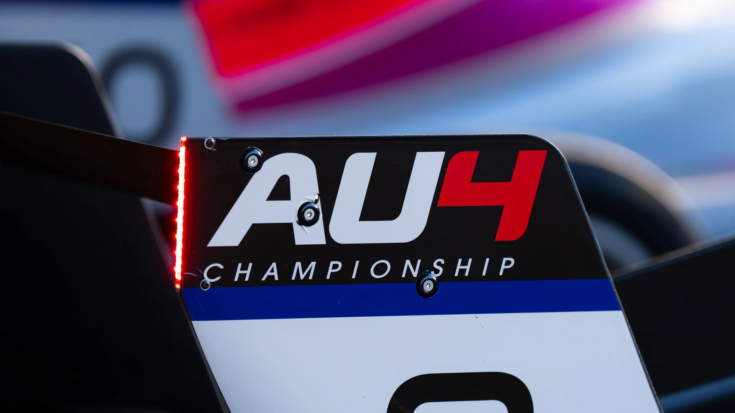 Close-up of a race car's rear wing with a 'AU4' championship logo and a red LED light.