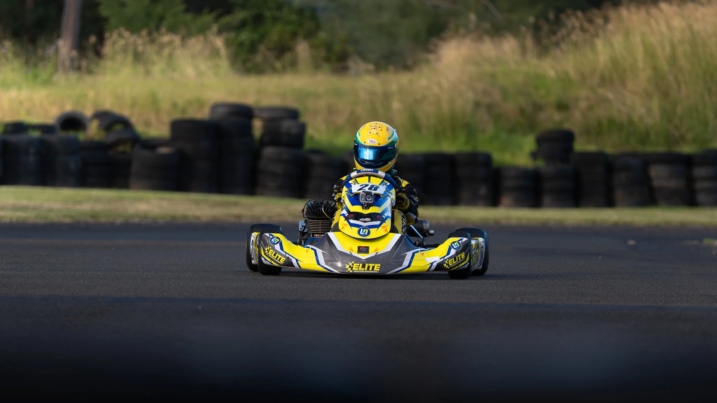 Go-kart race car with a yellow, black, and blue design on a racing track, driver wearing a matching helmet and racing suit.