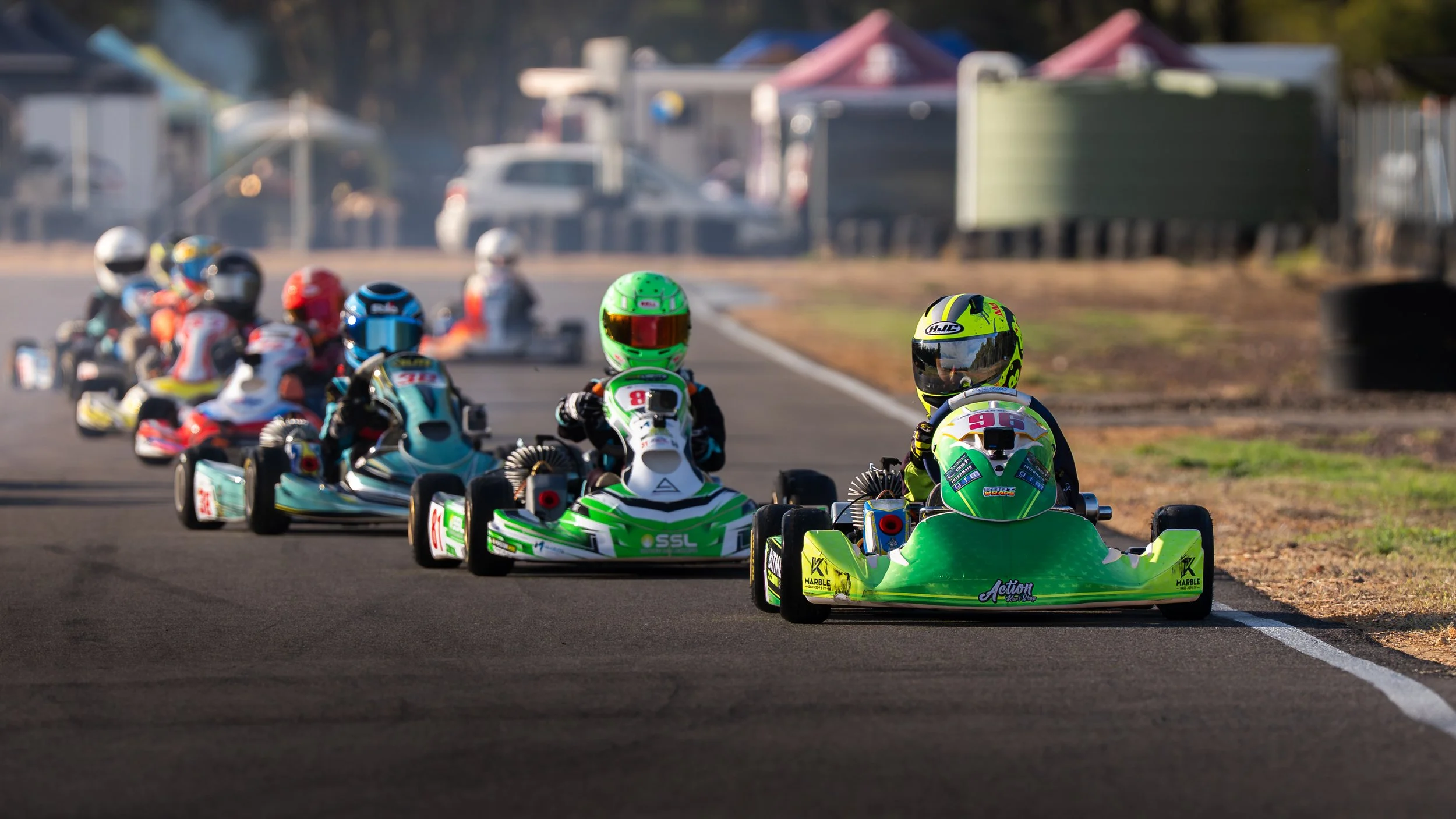 A group of go-kart racers in colorful helmets and racing suits on a race track, with the focus on two racers in the front yellow and green karts, as others follow behind.
