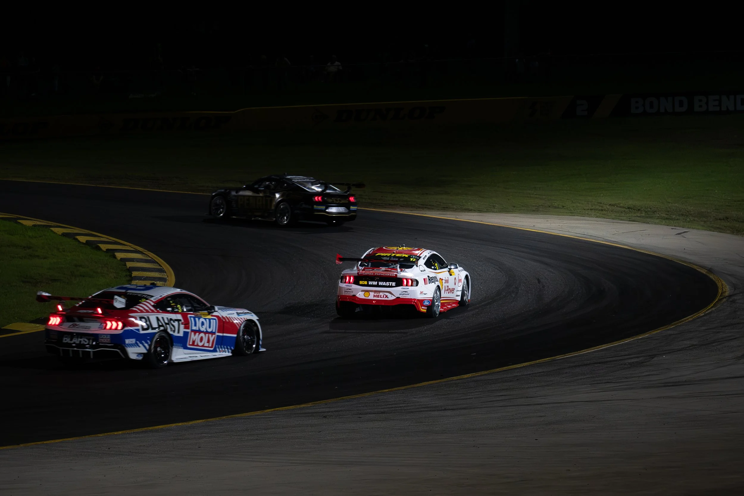 Nighttime Australian Supercars on a racetrack, including a white car with red and yellow logos and a black car, navigating a curve.