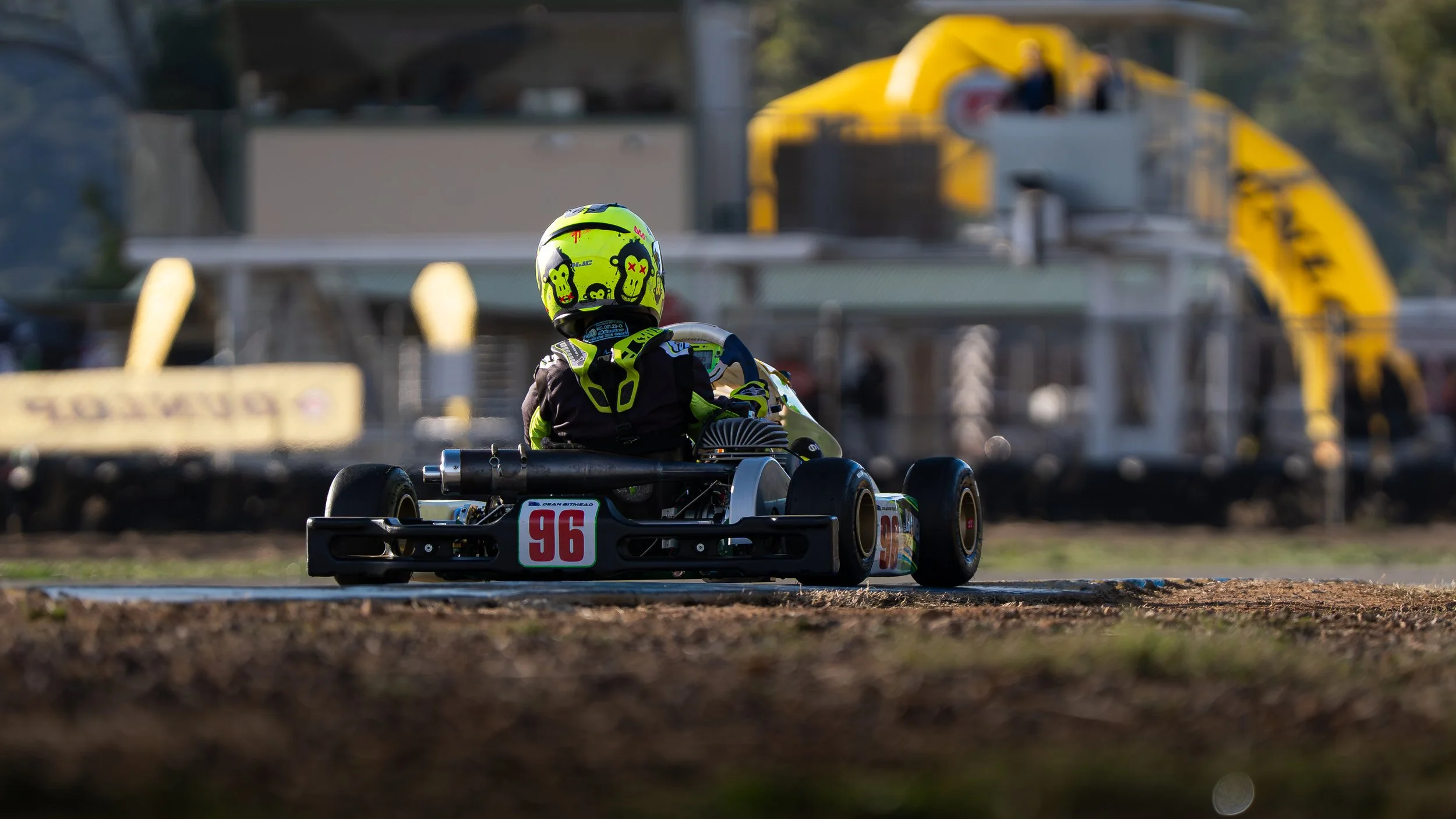 A person driving a go-kart on a dirt track with a racing helmet decorated with cartoon faces, wearing a black and yellow racing suit, number 96, and a yellow sign in the background.