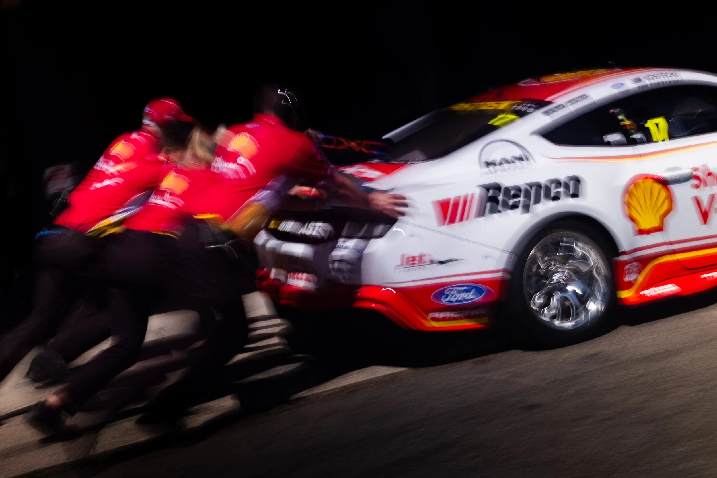 Race car crew members pushing a race car with sponsors' logos during a nighttime event.