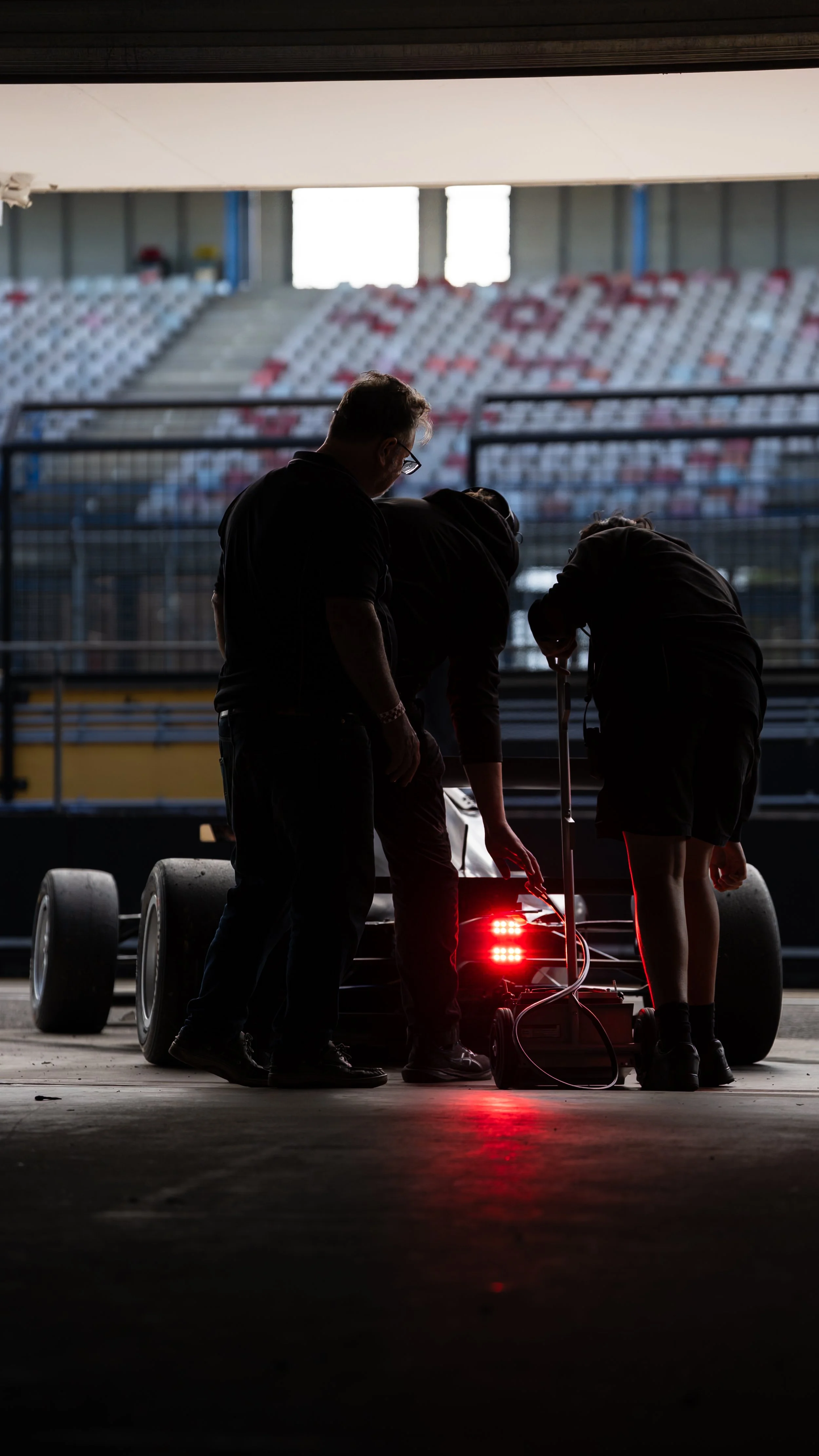 A group of people working on a race car in a garage, with the background of an empty stadium seating area visible through an opening.
